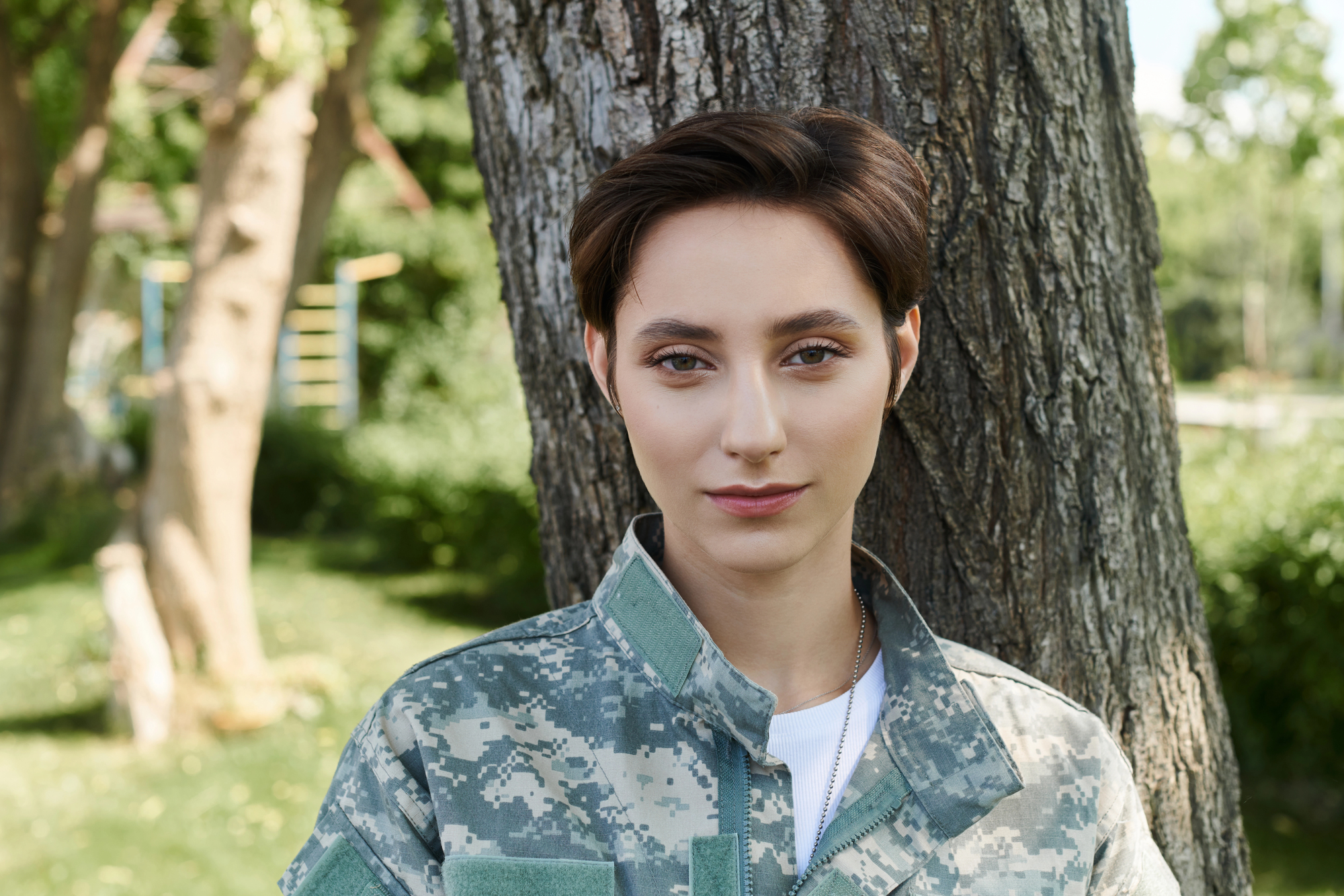 A person with short brown hair is leaning against a tree, wearing a camouflage-patterned military uniform. The background is lush with green trees and foliage, suggesting a park or outdoor setting. The individual is looking directly at the camera with a neutral expression.