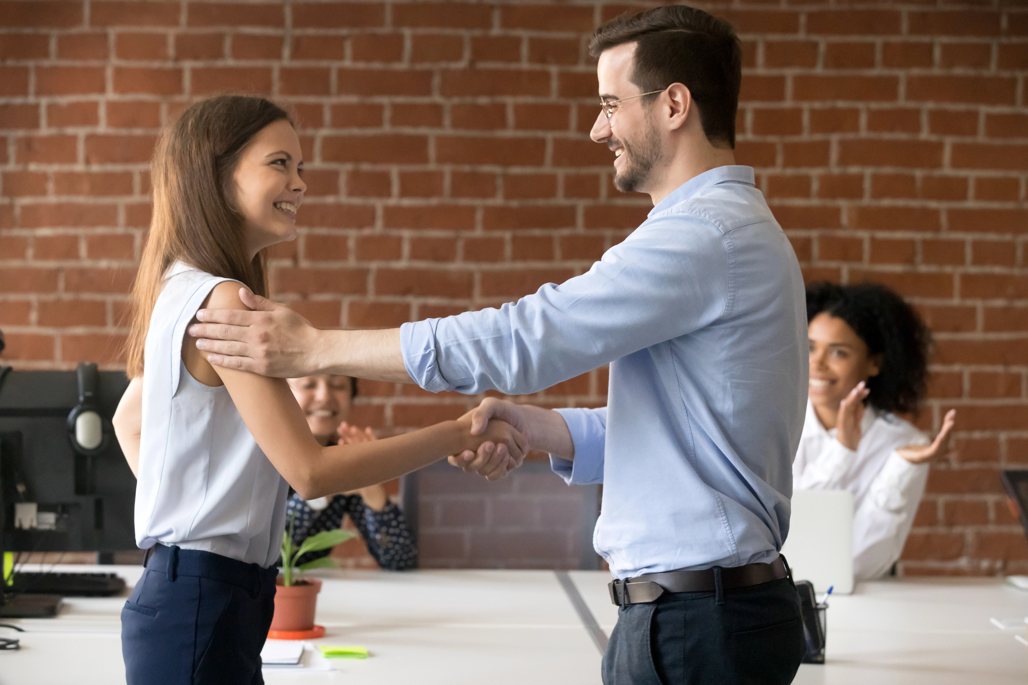 A man and a woman in business attire are shaking hands and smiling in an office with a brick wall. Another woman in the background is seated at a desk, clapping and smiling. An open laptop and a plant are also visible on the desks.