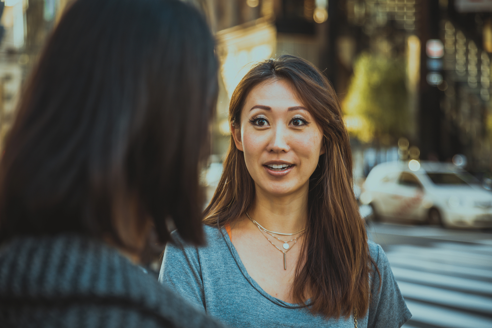 A woman with long brown hair and a gray shirt is engaged in conversation with another person whose back is to the camera. They are standing on a city street with blurred buildings and vehicles in the background, bathed in warm sunlight.