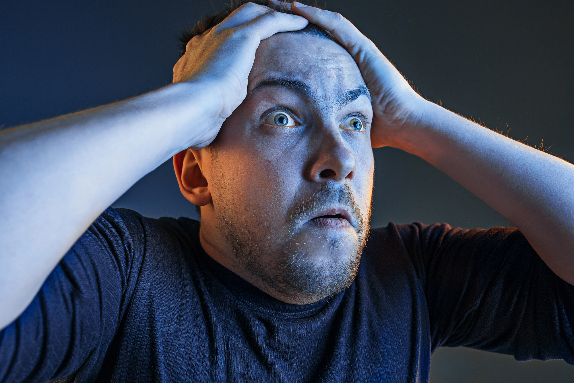 A man with a distressed expression holds his head with both hands. He has short hair, a beard, and is wearing a dark long-sleeve shirt. The background is dark, with dramatic lighting emphasizing his tense facial expression.