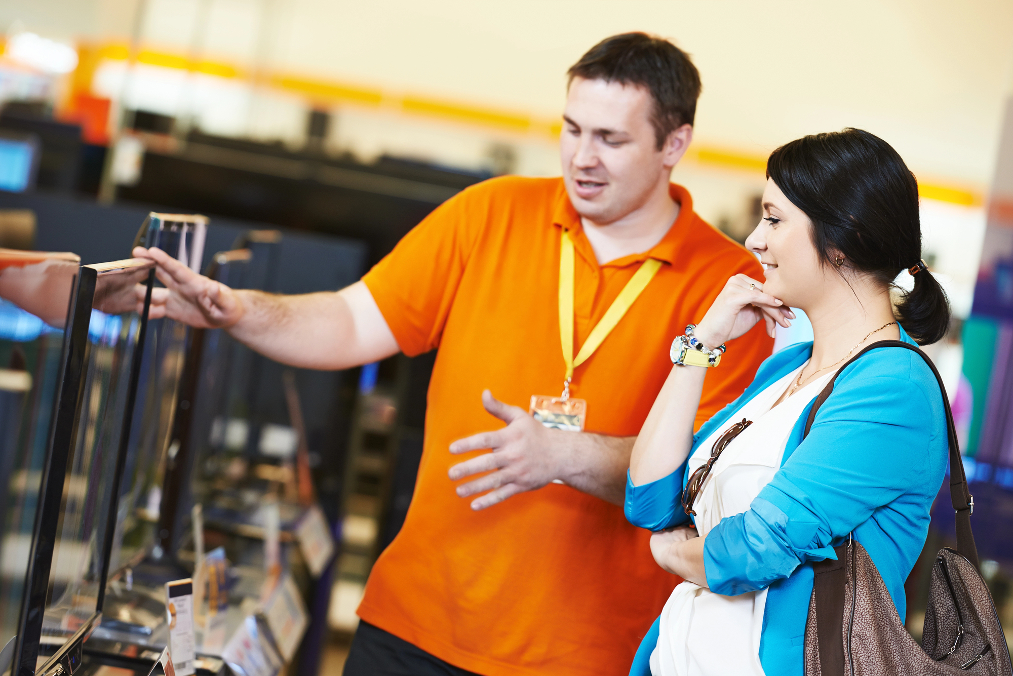 A store employee in an orange shirt with a name tag is assisting a customer wearing a blue cardigan. They are standing in front of a row of televisions, with the employee pointing at one of the screens while talking to the customer, who is listening attentively.