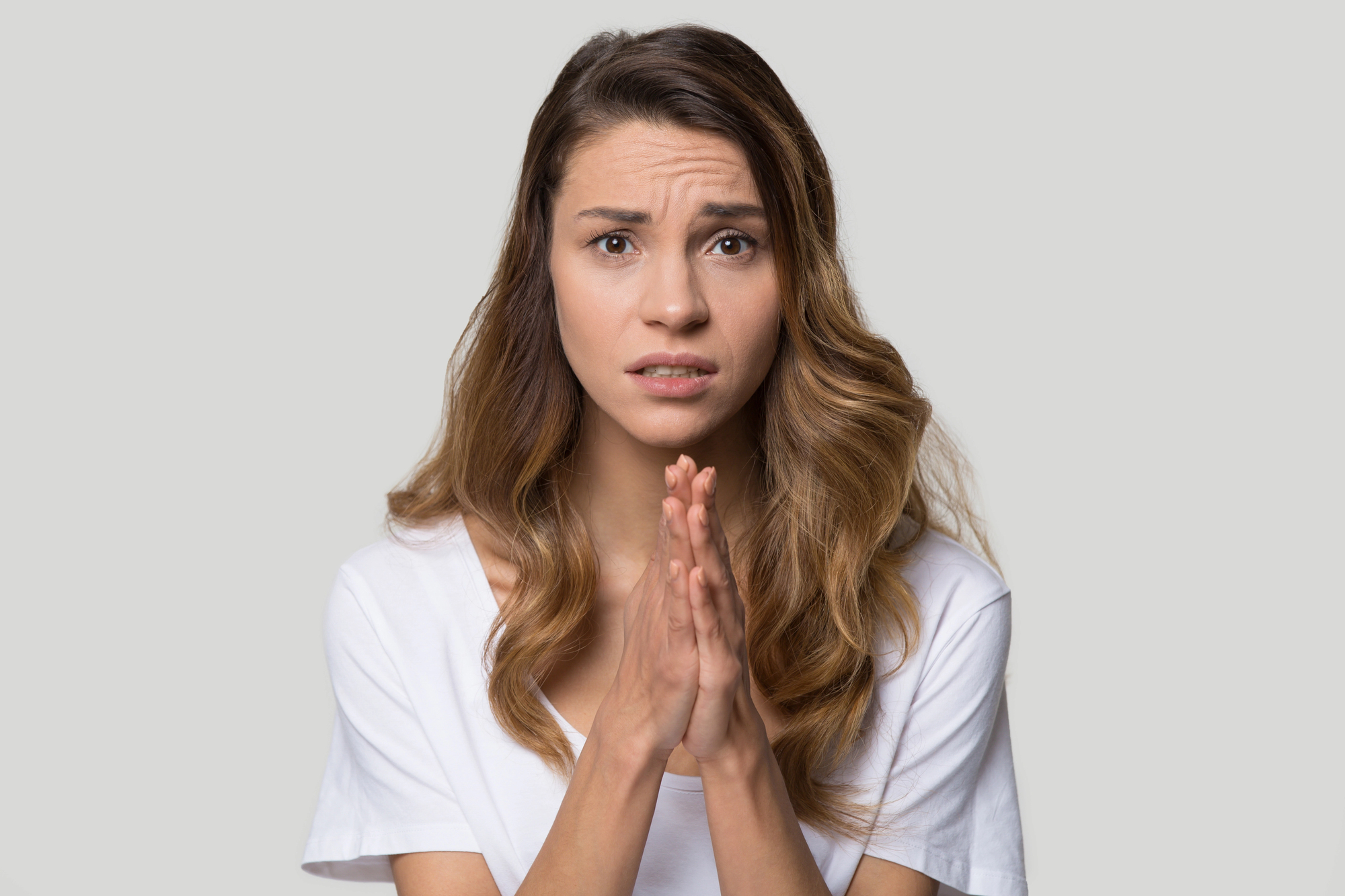 A woman with long, wavy, brown hair is looking at the camera with a worried expression. She is wearing a white shirt and clasping her hands together, as if in a pleading or praying gesture. The background is plain and light-colored.