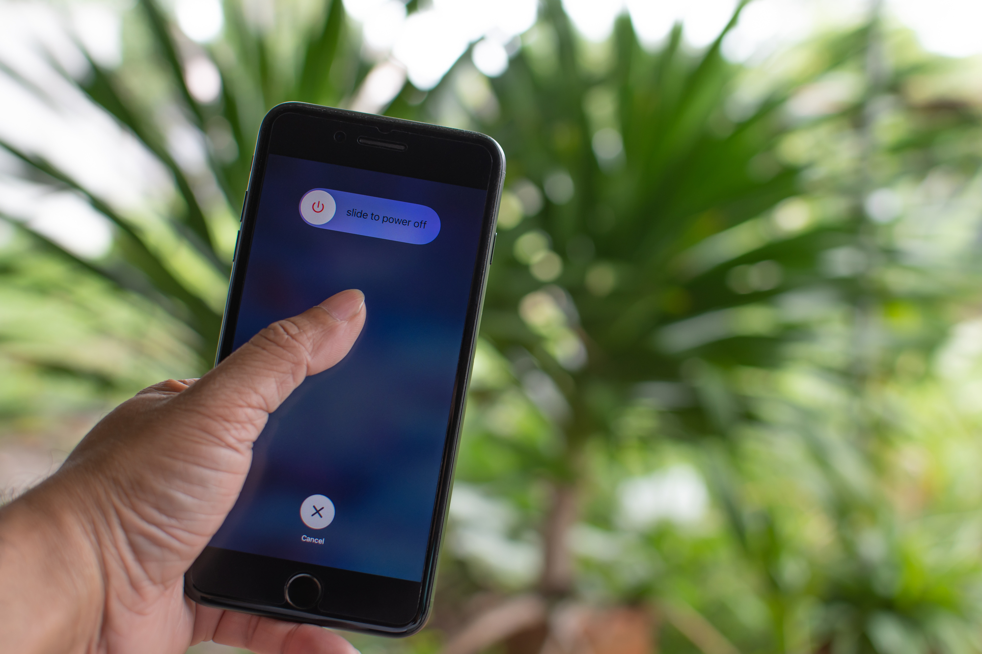 A hand holding a smartphone in front of greenery. The phone screen displays a "slide to power off" prompt with an on-screen power button and a cancel button at the bottom. The background is blurred, focusing on the phone and the hand.