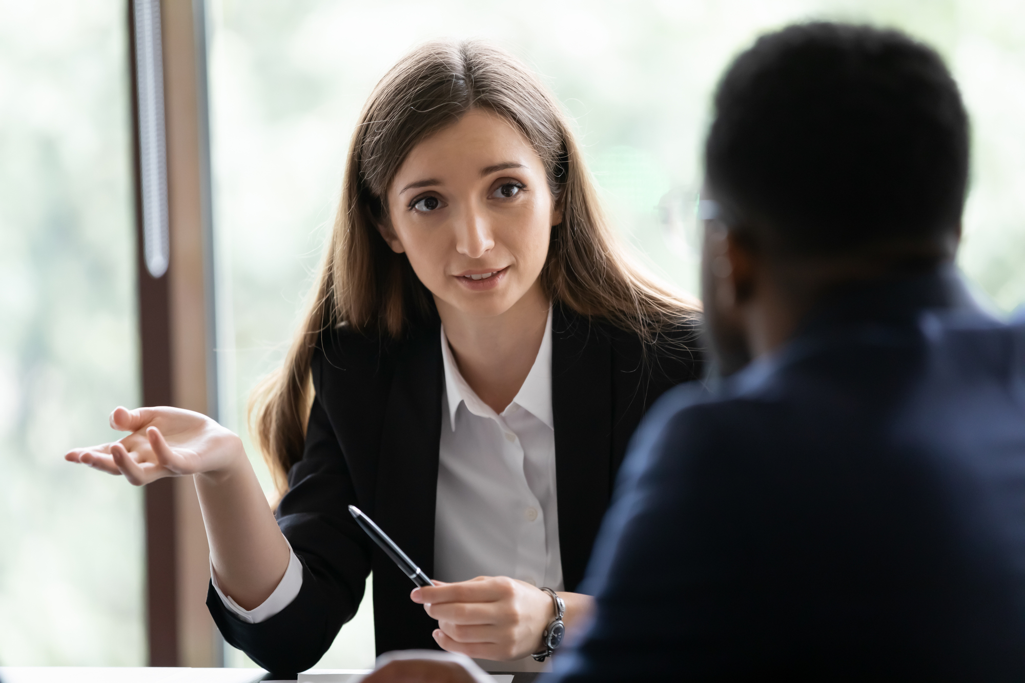 A woman with long brown hair, dressed in a black blazer, is conversing with a man in a dark blue suit. They are seated at a table with a bright, blurred background visible through large windows. The woman is gesturing with her right hand while holding a pen in her left hand.