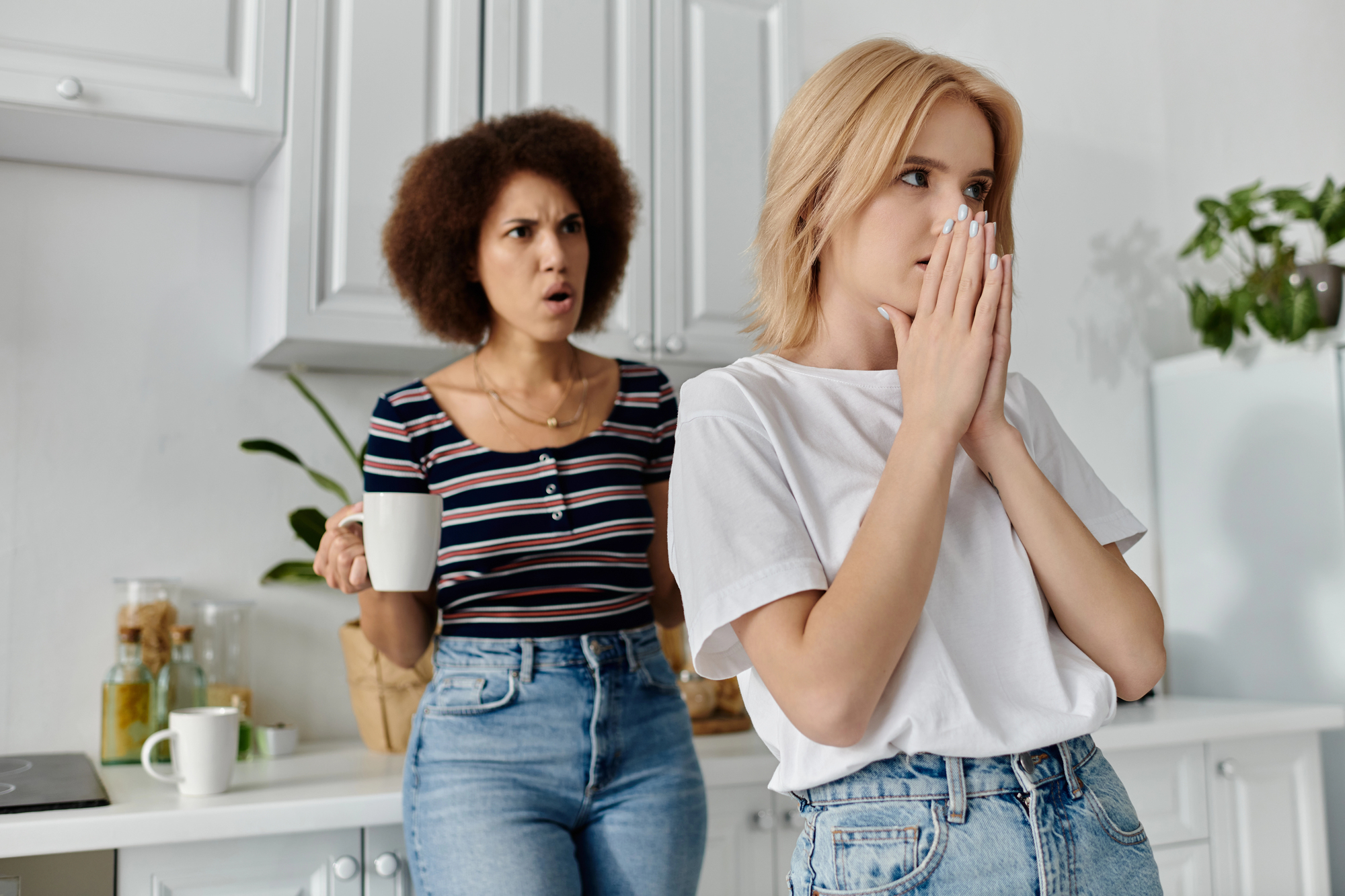 In a kitchen, two women appear to be having a disagreement. The woman in the foreground is looking away, covering her mouth with her hands, while the woman in the background is holding a mug and expressing surprise or frustration.