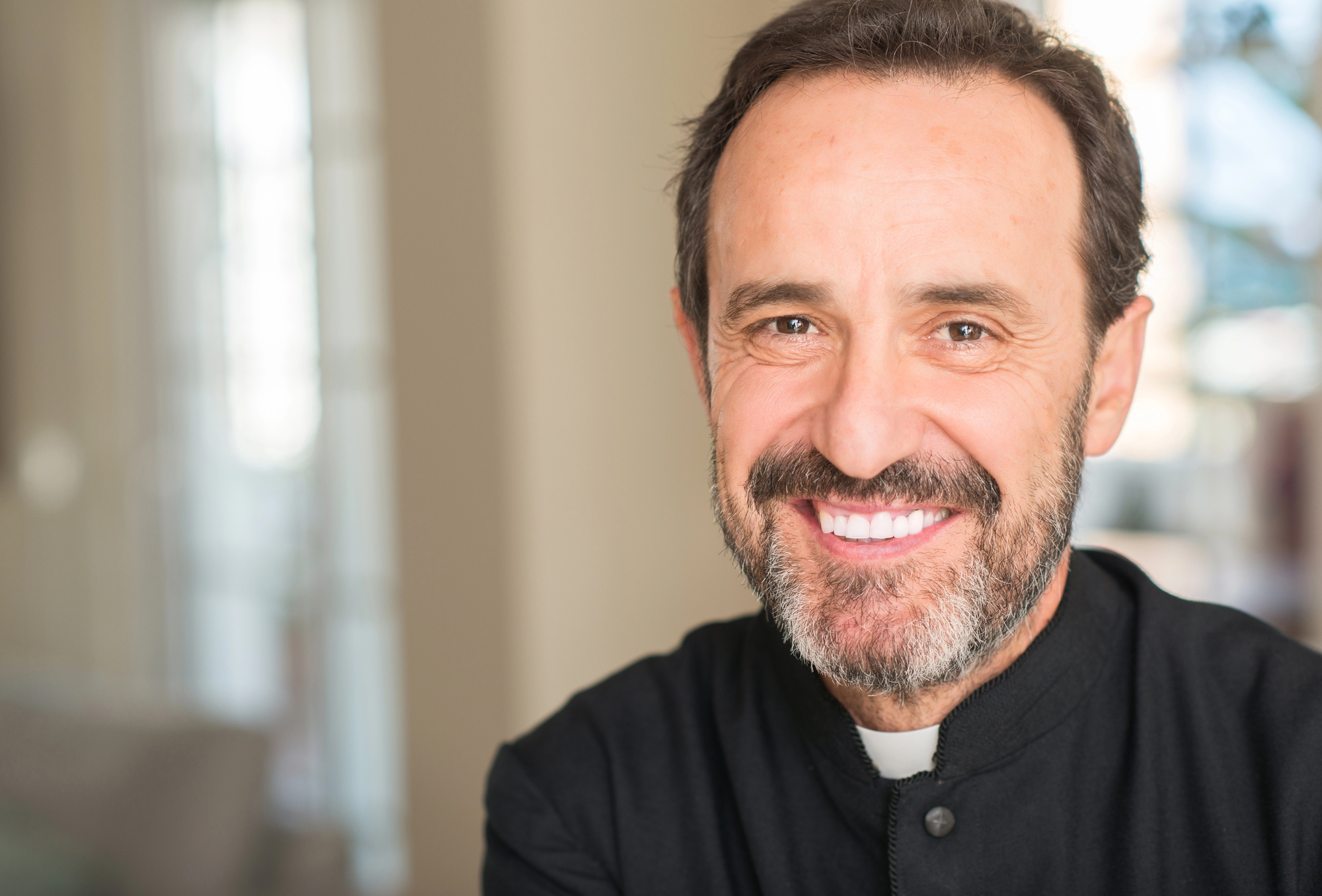 A middle-aged man with short dark hair and a beard smiles warmly at the camera. He is wearing a black clerical shirt with a white collar. The background is softly blurred, featuring bright, natural light coming through windows.