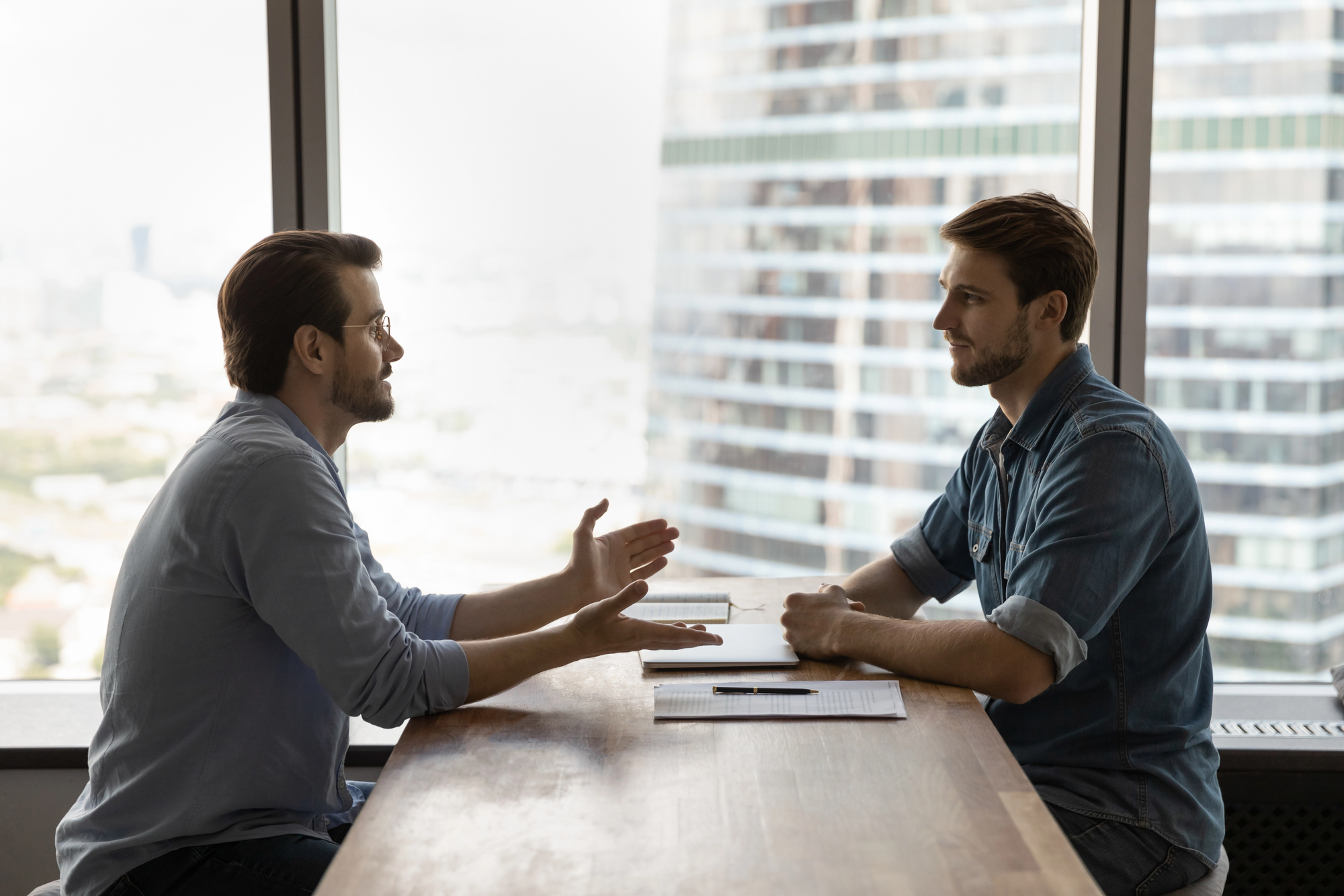 Two men are sitting across from each other at a long wooden table in a modern office with cityscape windows. They appear to be having a serious conversation. One man is gesturing with his hands, while the other listens attentively.