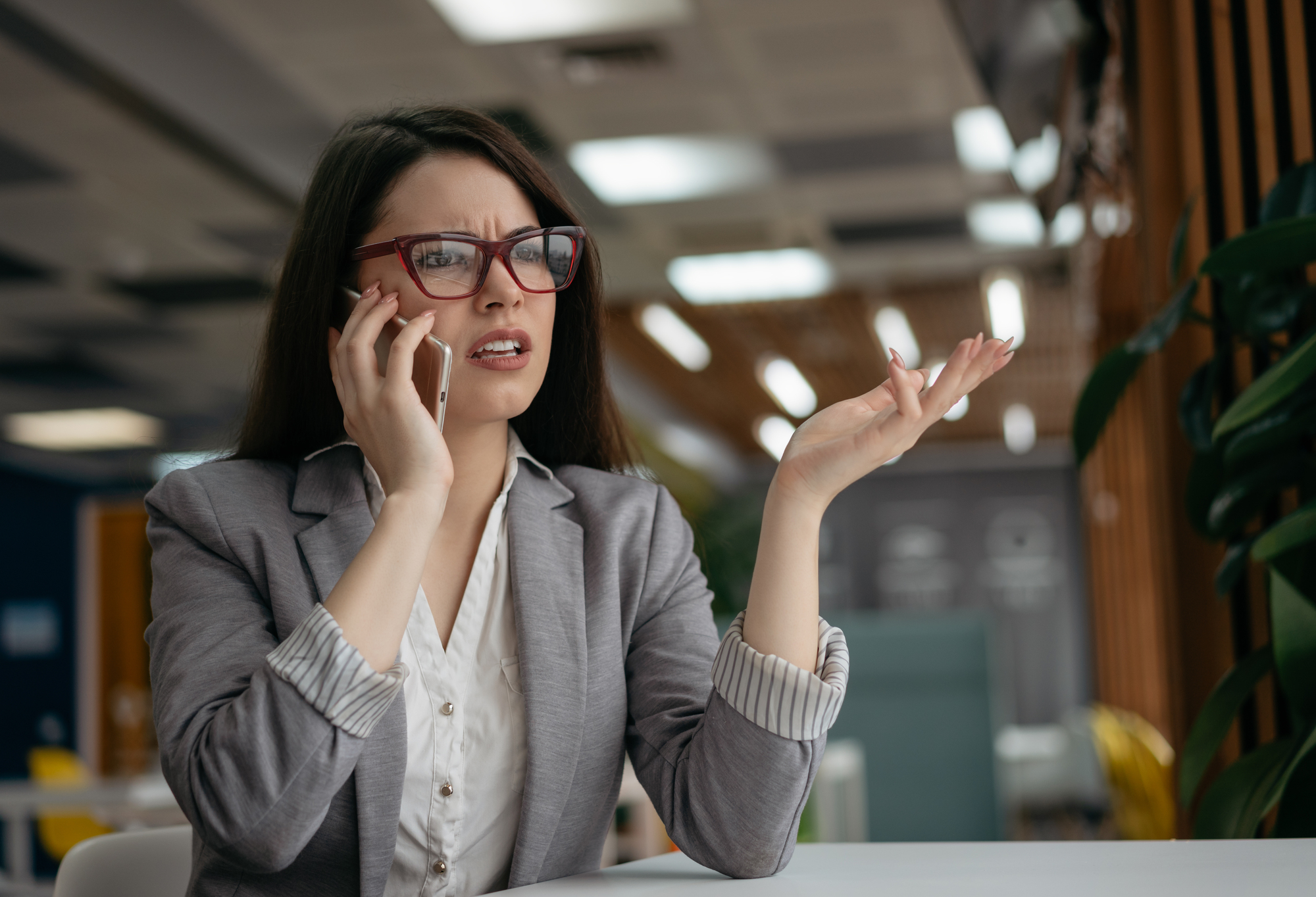 A woman with long dark hair and glasses is sitting at a desk in an office. She is wearing a gray blazer and white shirt and appears frustrated while speaking on her mobile phone. Her right hand is raised in an exasperated gesture.