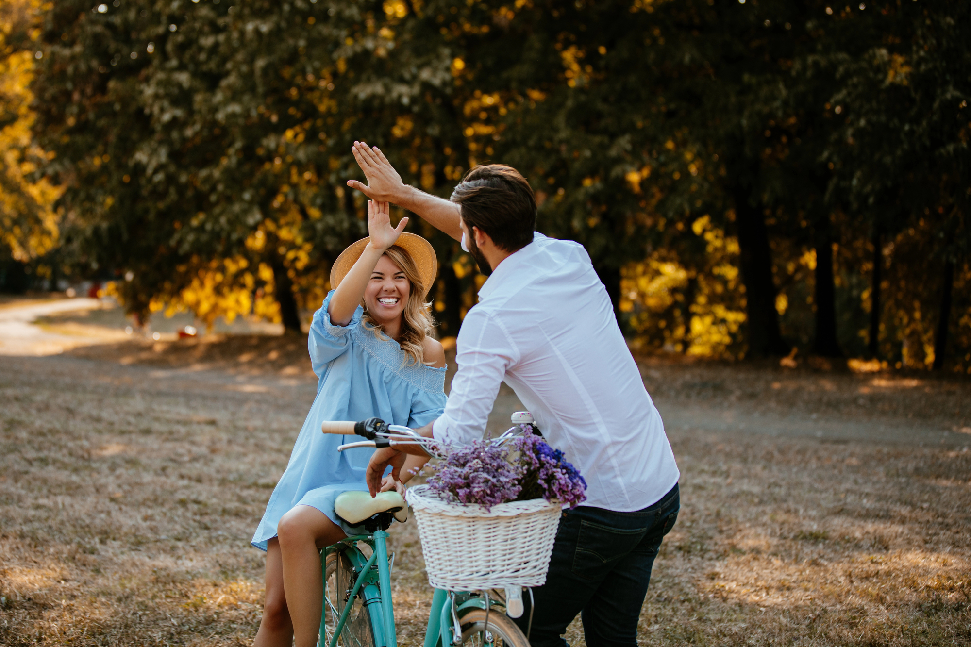 A woman sitting on a bicycle with a basket of purple flowers high-fives a man standing next to her in a park. The woman is wearing a blue dress and a wide-brimmed hat, while the man is dressed in a white shirt. Trees with sunlight filtering through the leaves are in the background.