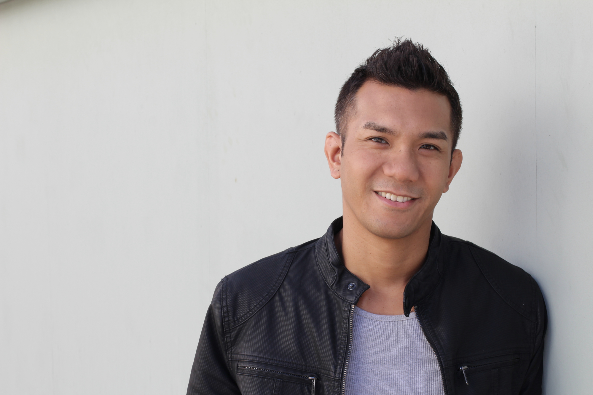 A man with short dark hair stands against a light-colored wall. He is wearing a dark leather jacket over a gray shirt and is smiling at the camera.