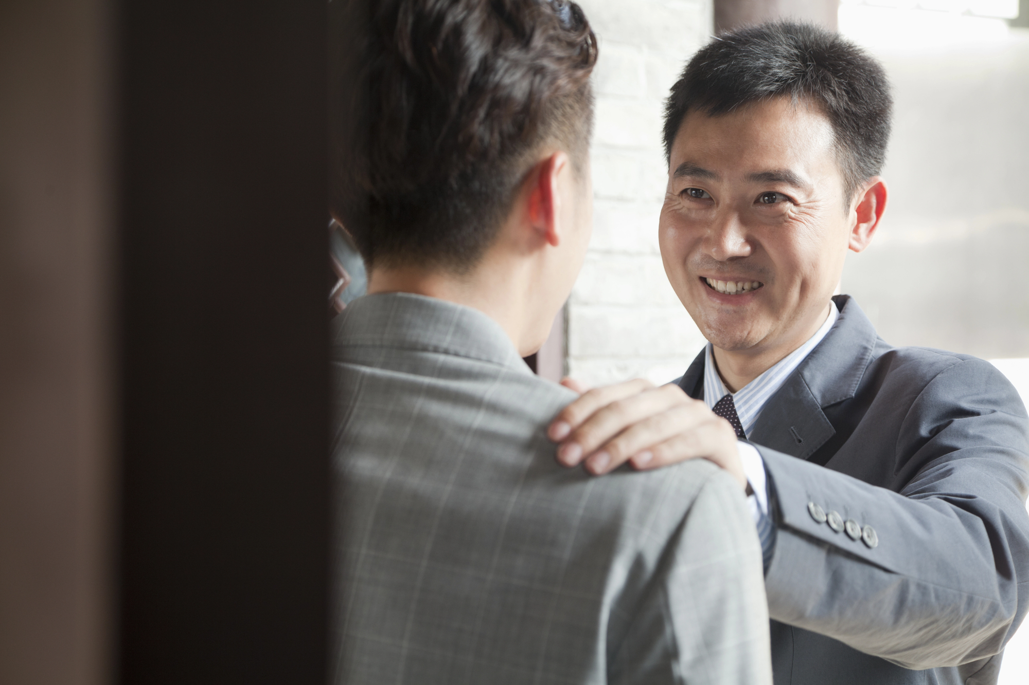 A man in a suit smiles and places his hand on the shoulder of another man who is turned away from the camera and also wearing a suit. They are in an indoor setting with natural light coming through a window.