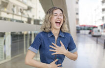 A person with shoulder-length wavy hair laughs heartily while holding their chest and stomach. They are wearing a short-sleeved denim dress and standing on a city street with buildings and a bus in the background.