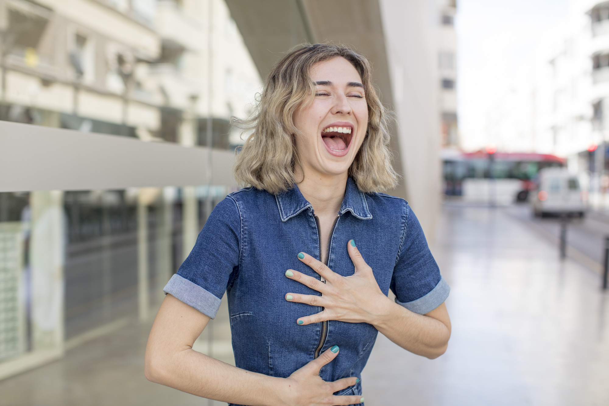 A person with shoulder-length wavy hair laughs heartily while holding their chest and stomach. They are wearing a short-sleeved denim dress and standing on a city street with buildings and a bus in the background.