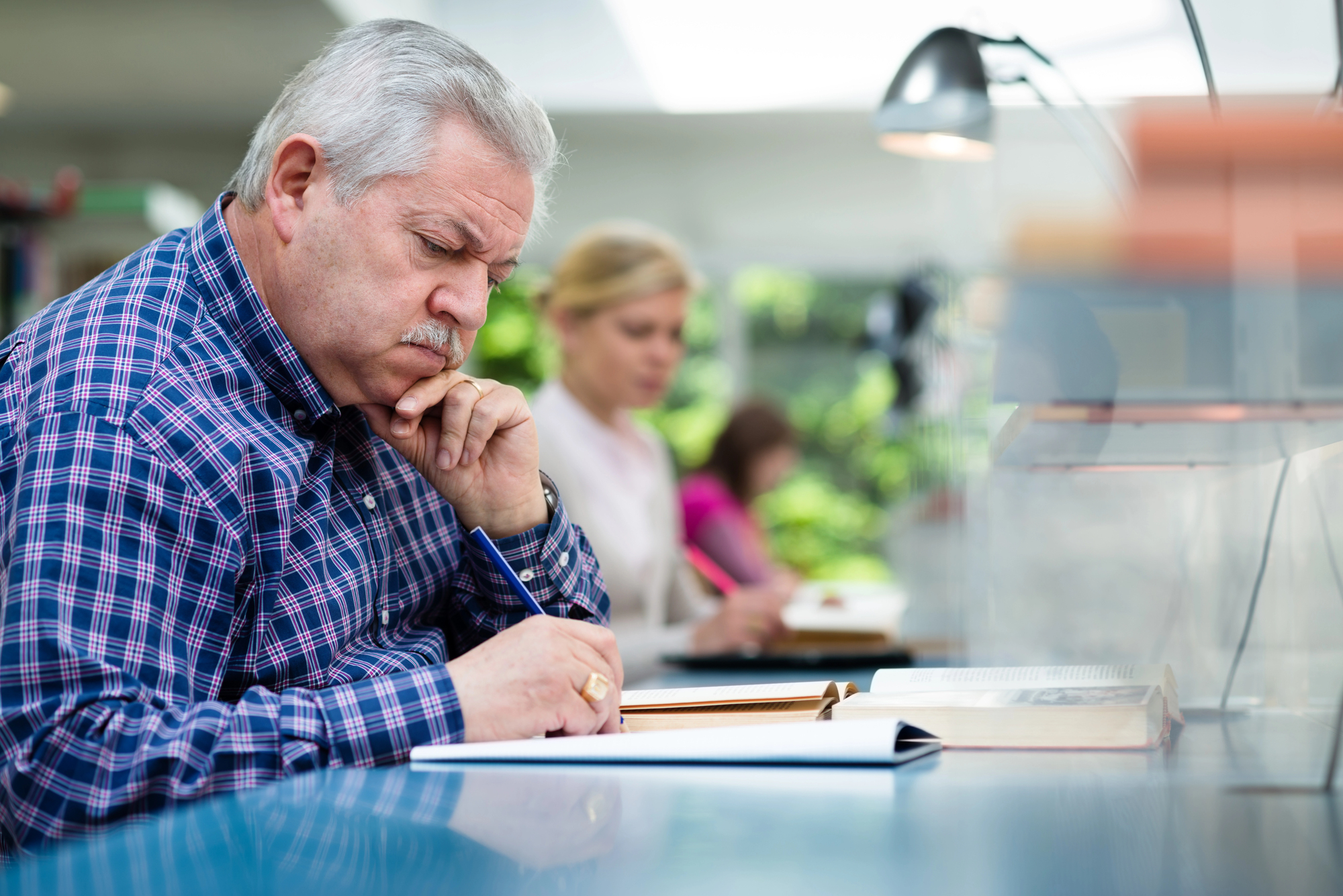 An older man with gray hair, wearing a blue and red plaid shirt, is sitting at a desk in a library, intently reading a book and taking notes. In the background, other individuals are also reading at the desks. The atmosphere appears quiet and studious.