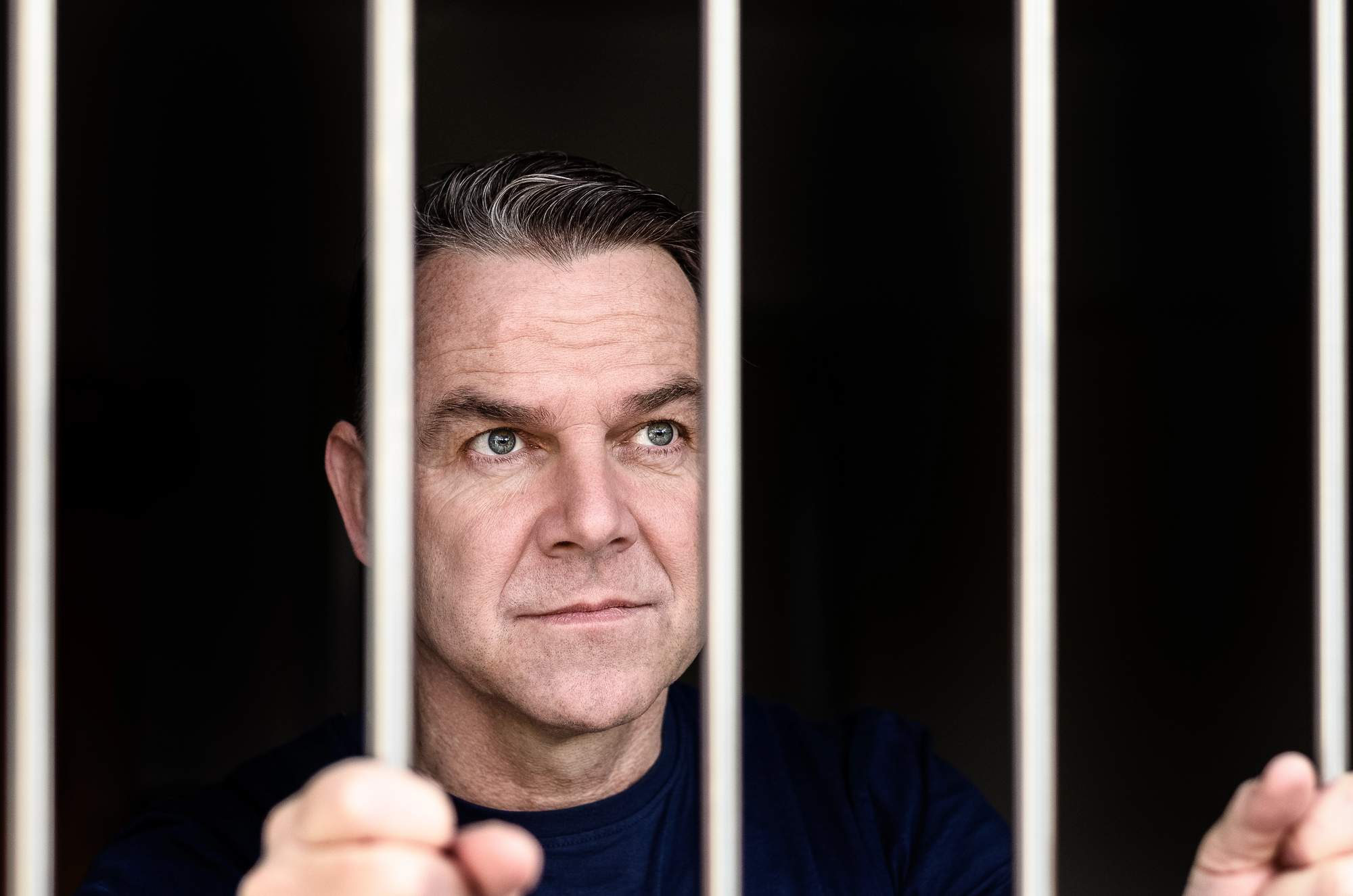 A man with short, dark hair holds onto metal bars, looking pensively through them. The background is dark, emphasizing the feeling of confinement.