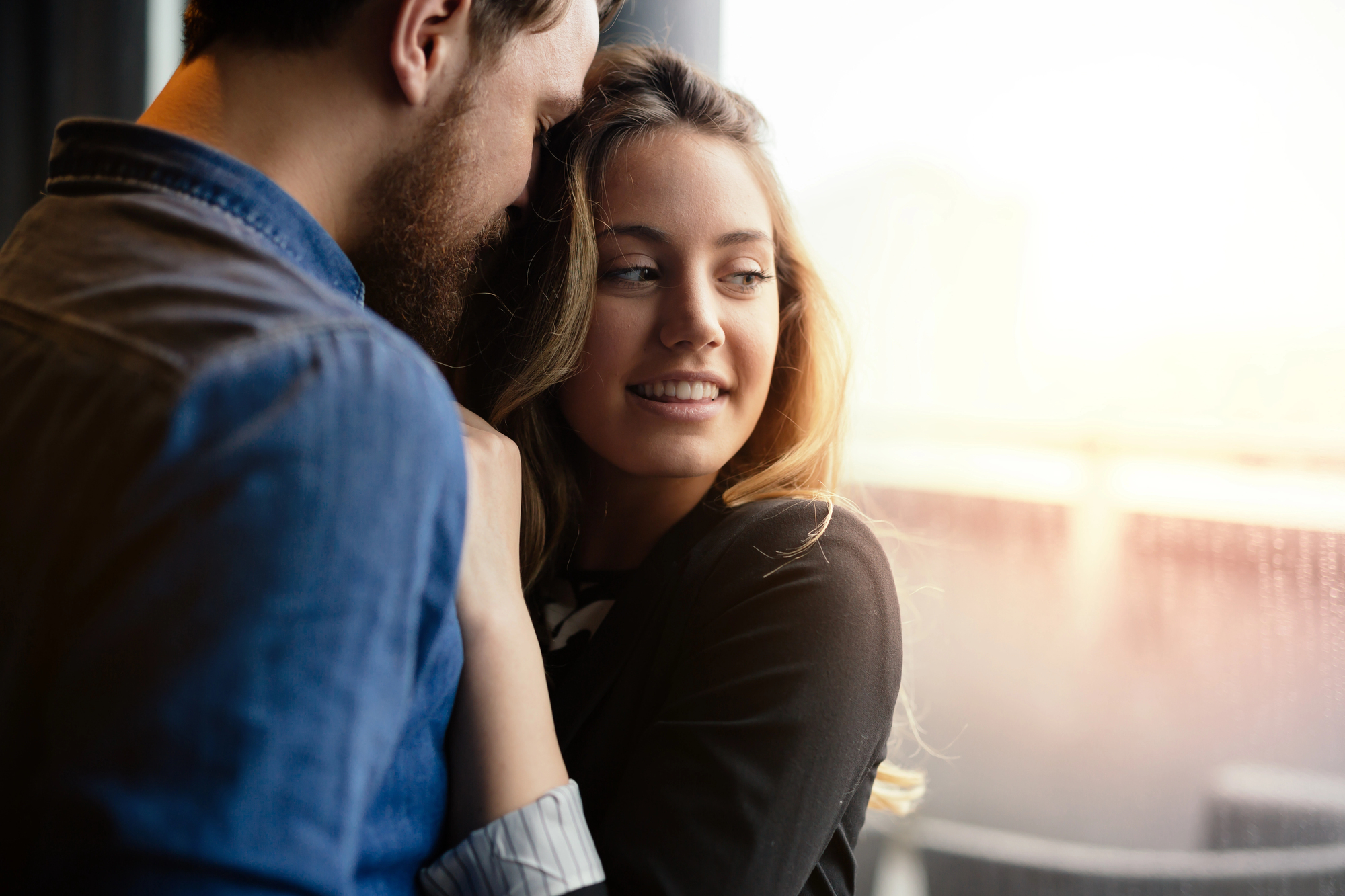 A young couple stands close to each other by a window. The man has a beard and is whispering or speaking softly to the woman, who is smiling and looking out the window. Sunlight streams through the window, creating a warm, glowing atmosphere.