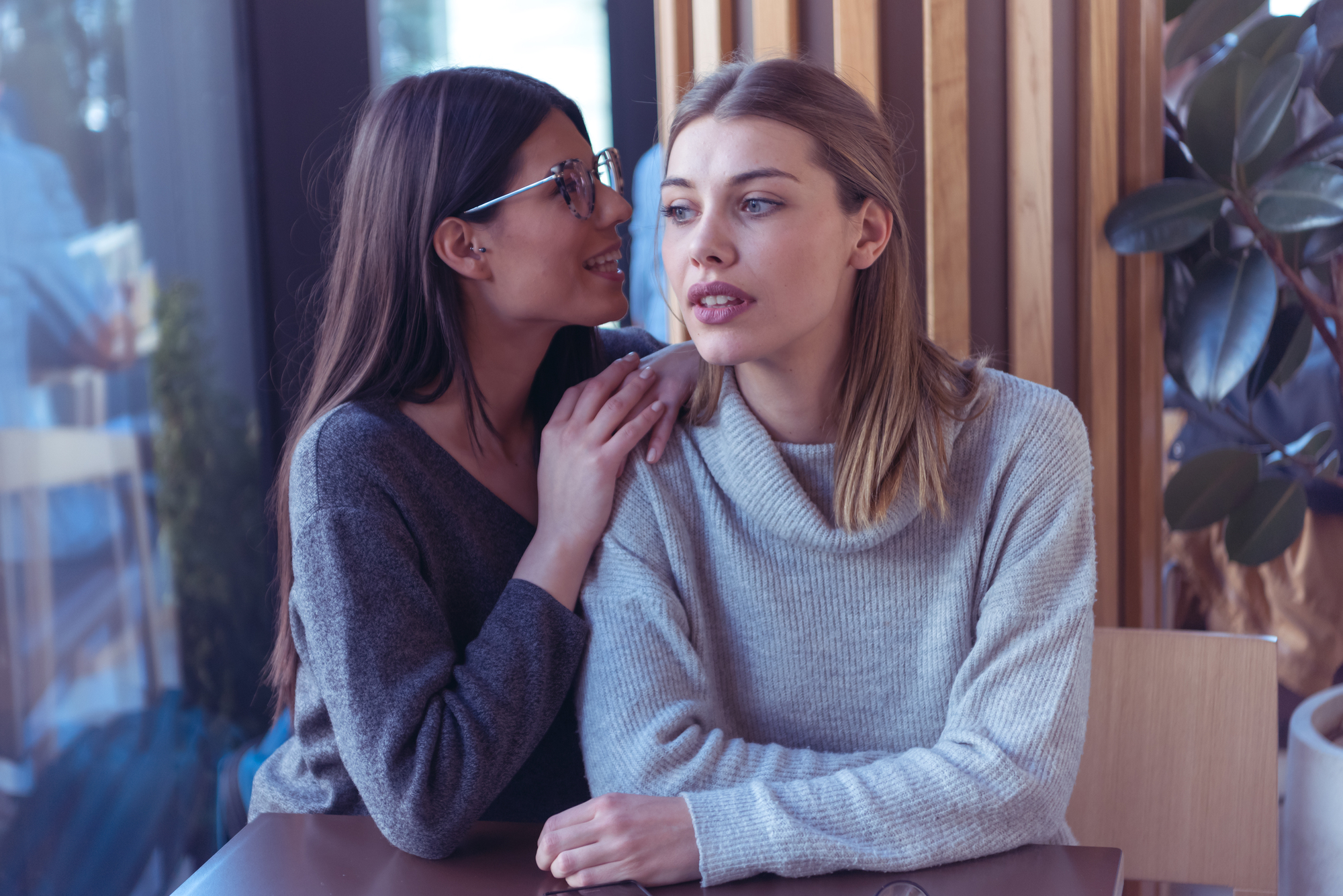 Two women sit at a table; one leans in to whisper into the other's ear, with a hand resting on her shoulder. The listener looks forward with a neutral expression. They are indoors, and wooden panels and a plant are partially visible in the background.