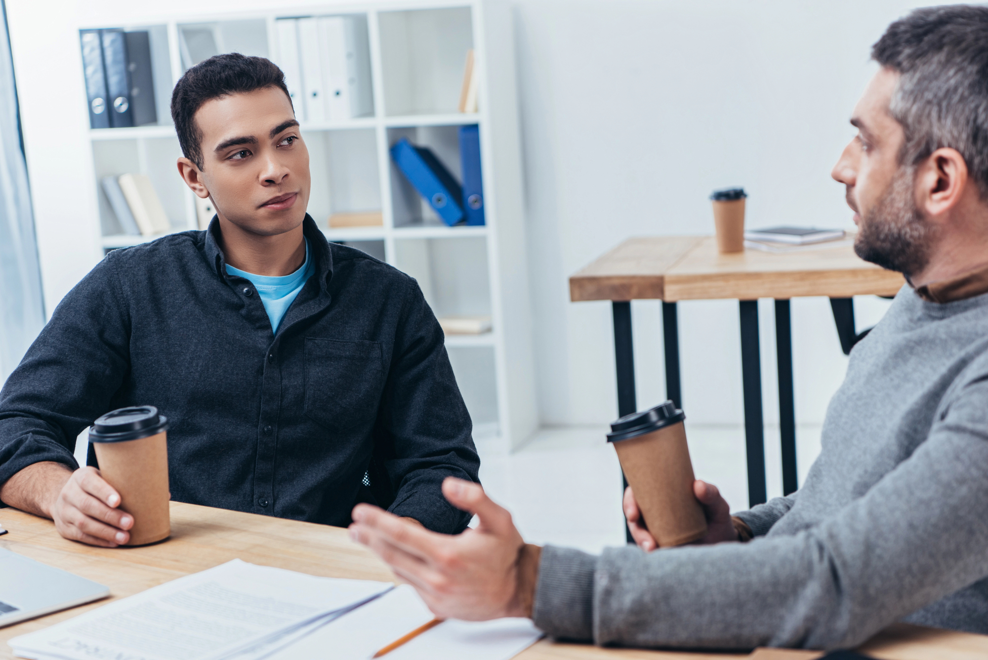 Two men are having a conversation over coffee in a modern office setting. Both are sitting at a wooden table with documents and papers on it. One man gestures with his hand while talking, and the other listens attentively. Bookshelves and desks are visible in the background.