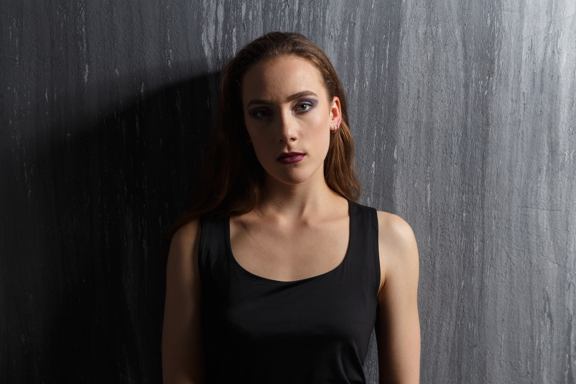 A woman with long brown hair and dark lipstick stands against a dark, textured wall. She wears a sleeveless black top and has a serious expression on her face. The lighting is low, casting shadows that add to the moody atmosphere.