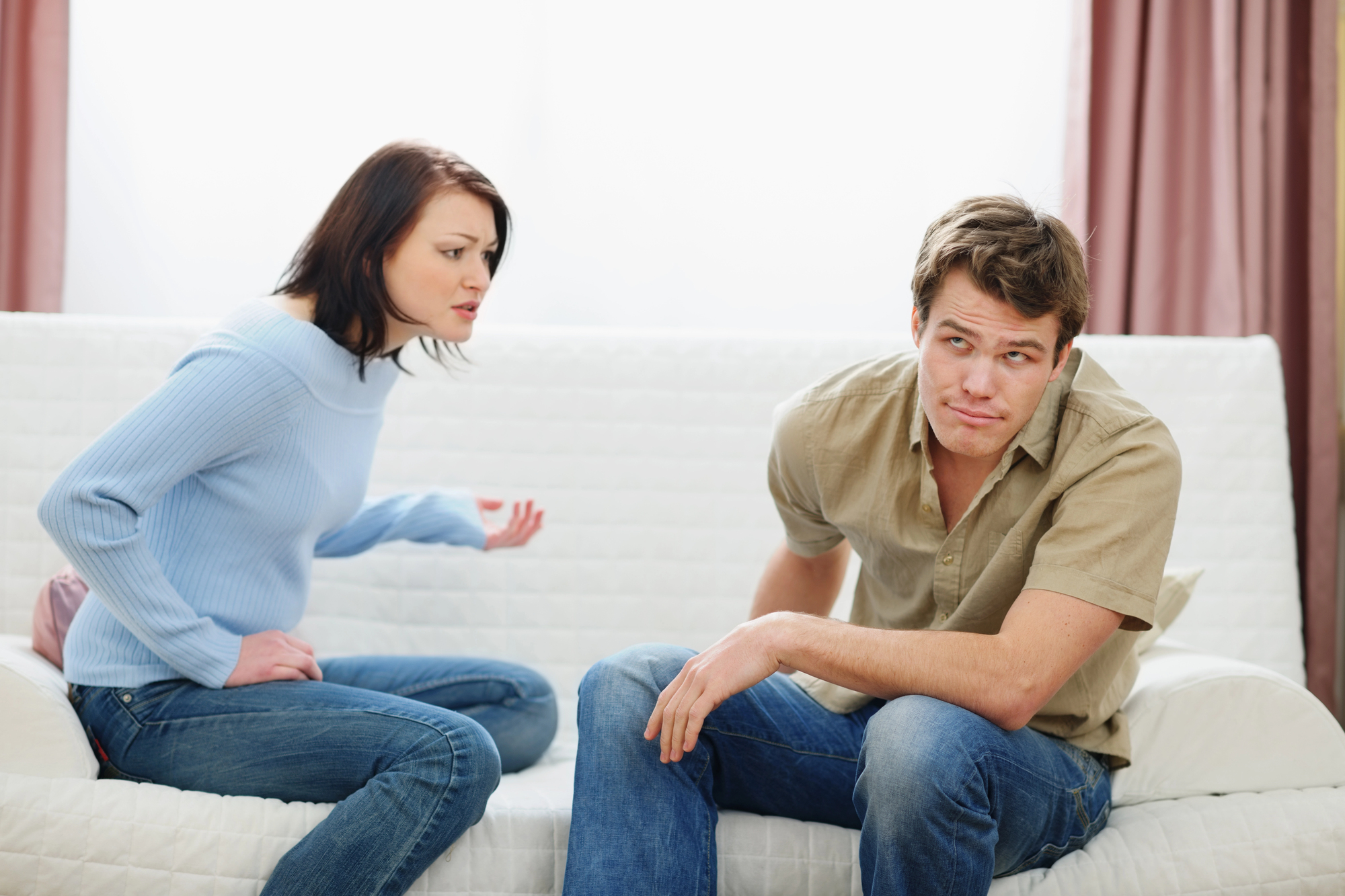 A woman in a light blue sweater appears to be confronting a man in a beige shirt who sits next to her on a white couch. The woman is gesturing with her hand while the man looks away with a slightly frustrated expression.