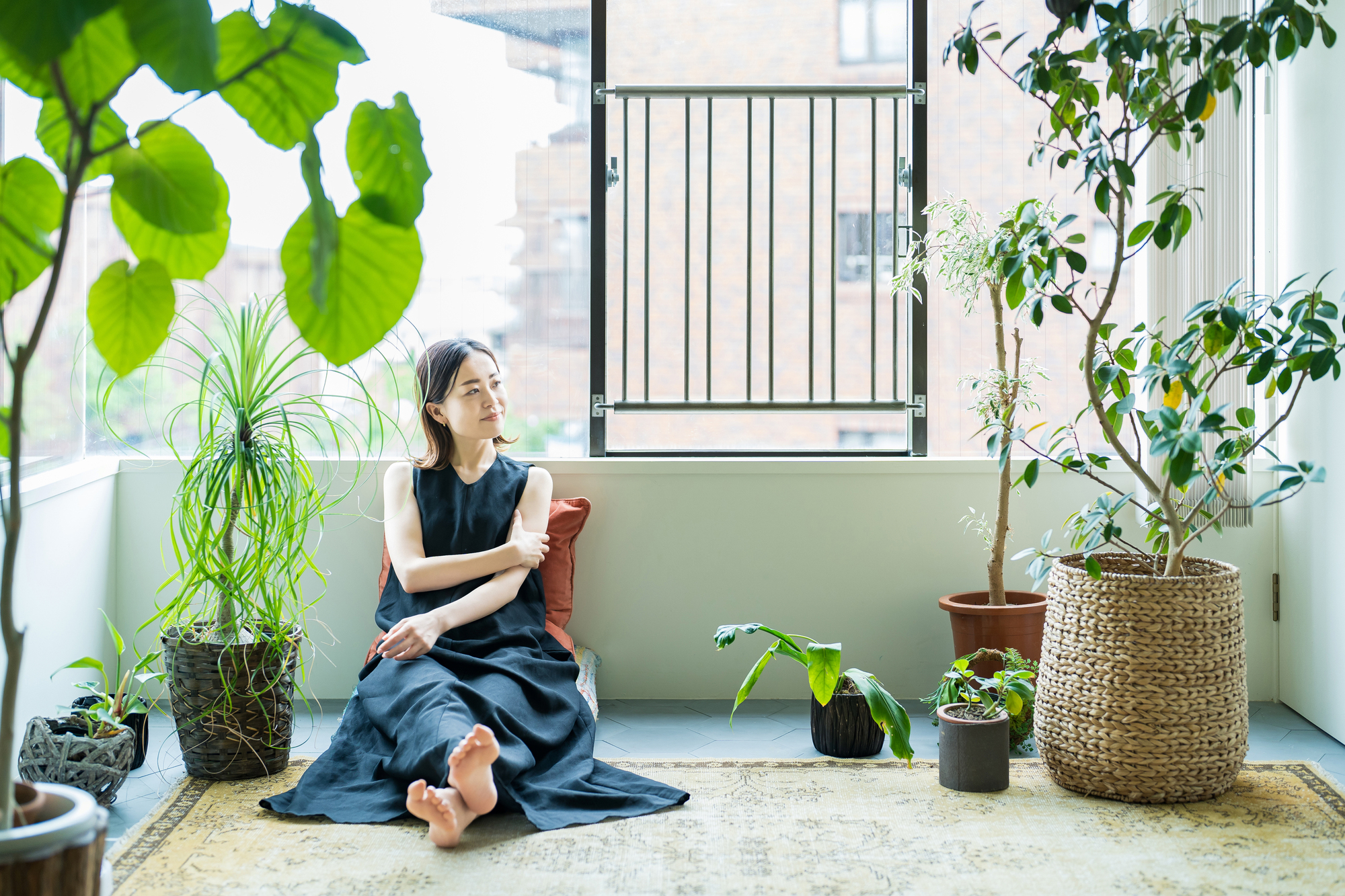 A woman in a black dress sits on the floor near a window surrounded by various potted plants. She rests her head on her hand, looking thoughtfully out the window. The room is bright with natural light, and a rug covers the floor.