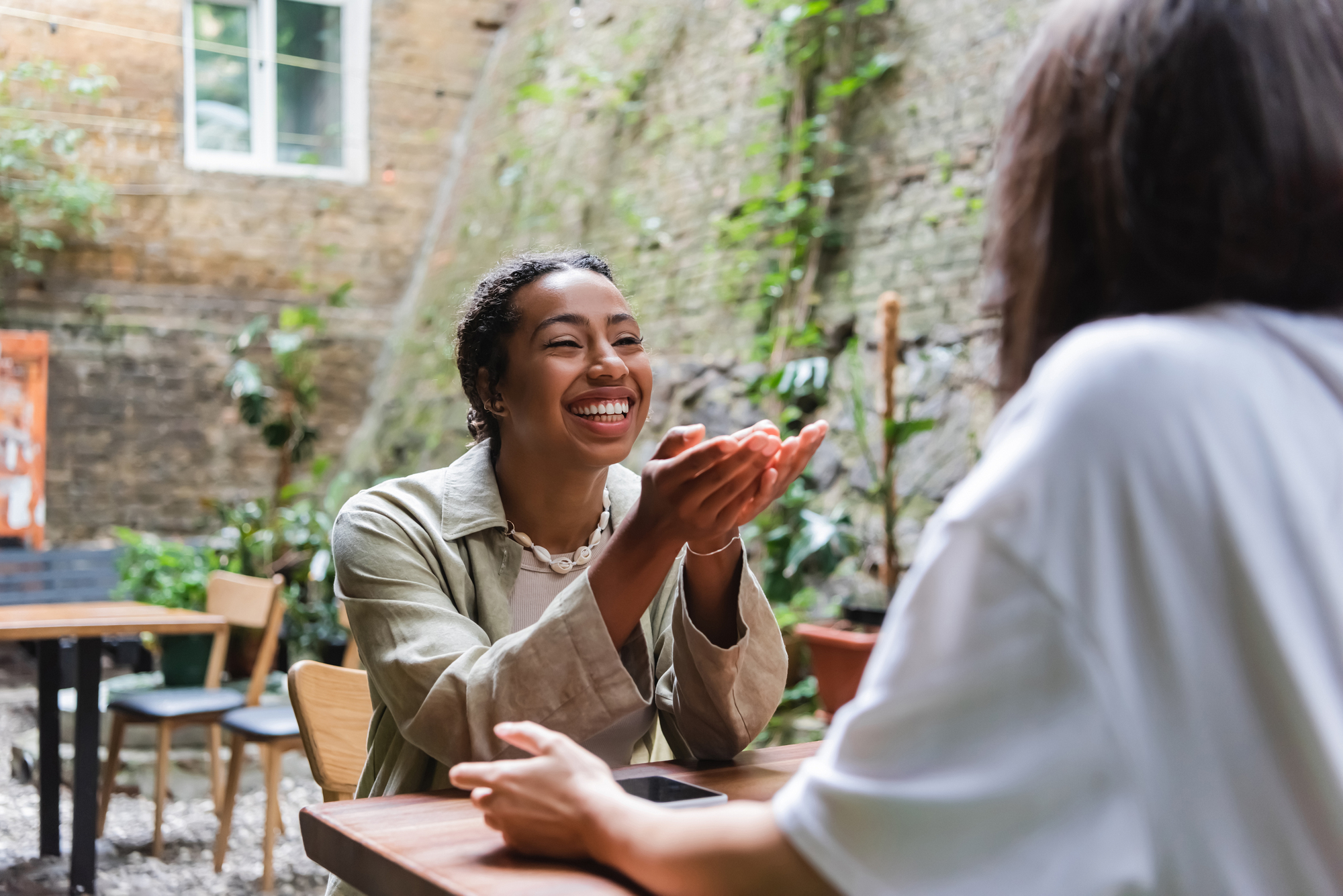 Two people are sitting at an outdoor café. One person, facing the camera, is smiling and speaking while gesturing with their hands. The other person has their back to the camera and is listening attentively. The background features greenery and a brick wall.