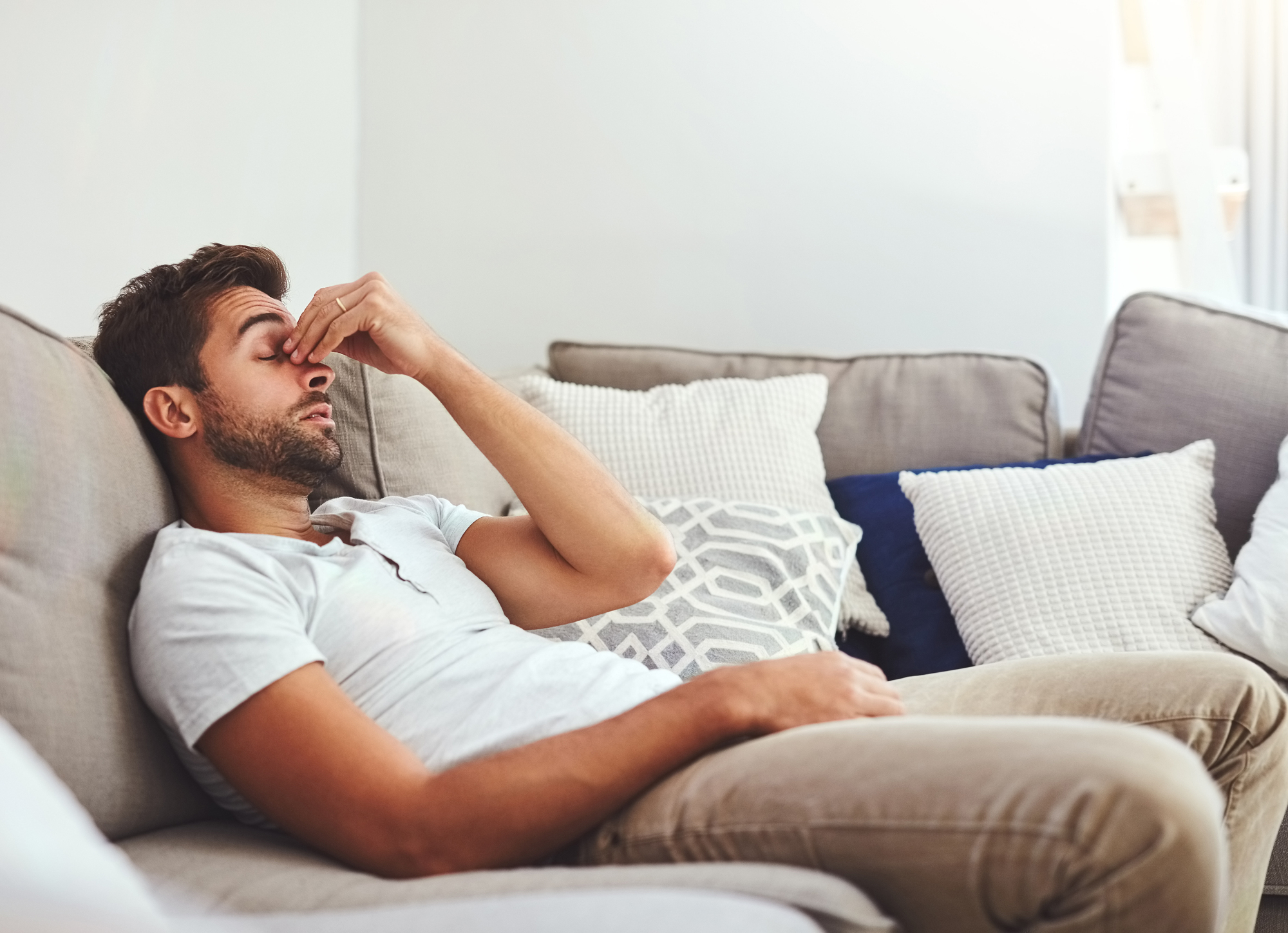 A man with a beard is lying on a couch, wearing a light-colored t-shirt and beige pants. He has his eyes closed and is pinching the bridge of his nose, appearing to be stressed or exhausted. The couch is decorated with several patterned and plain cushions.