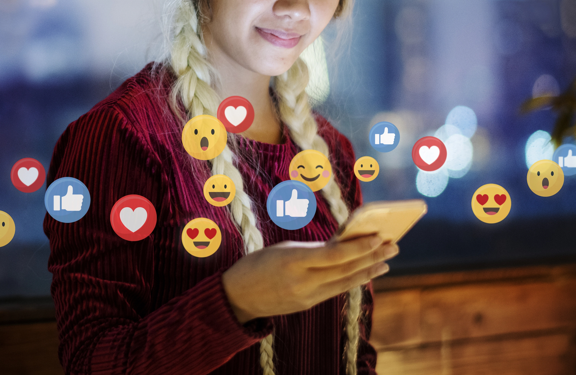 A person with light hair in braids is holding a smartphone and smiling slightly. Various social media reaction icons, such as thumbs up, hearts, and smiley faces, are overlaid around the person, indicating engagement with content on their device.