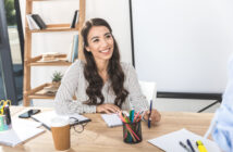 A woman with long dark hair, wearing a striped blouse, is sitting at a desk and smiling. The desk has a notebook, colorful pens, a take-out coffee cup, and various office supplies. In the background, there is a wooden shelf with books and a potted plant.