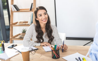 A woman with long dark hair, wearing a striped blouse, is sitting at a desk and smiling. The desk has a notebook, colorful pens, a take-out coffee cup, and various office supplies. In the background, there is a wooden shelf with books and a potted plant.