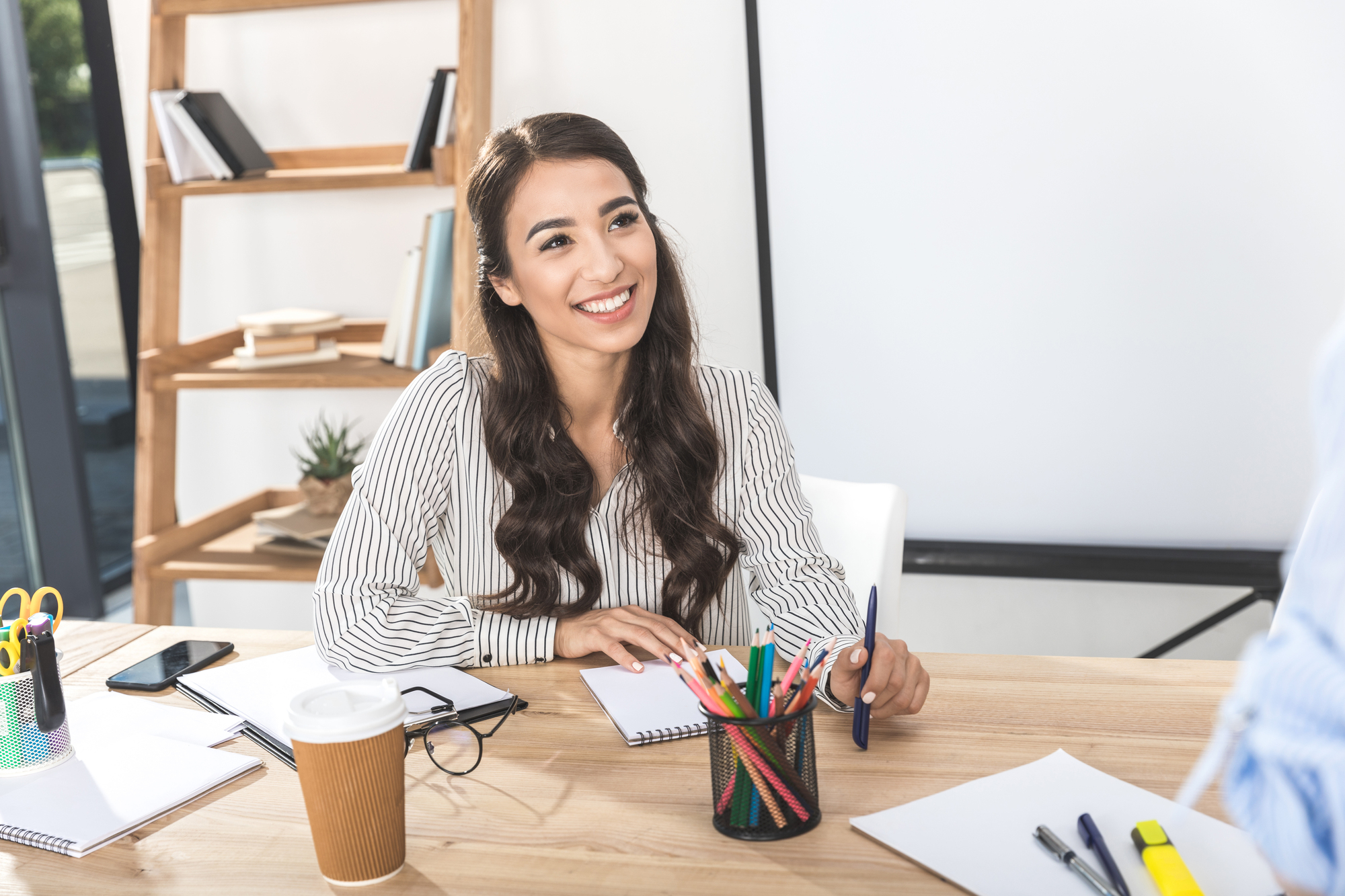 A woman with long dark hair, wearing a striped blouse, is sitting at a desk and smiling. The desk has a notebook, colorful pens, a take-out coffee cup, and various office supplies. In the background, there is a wooden shelf with books and a potted plant.