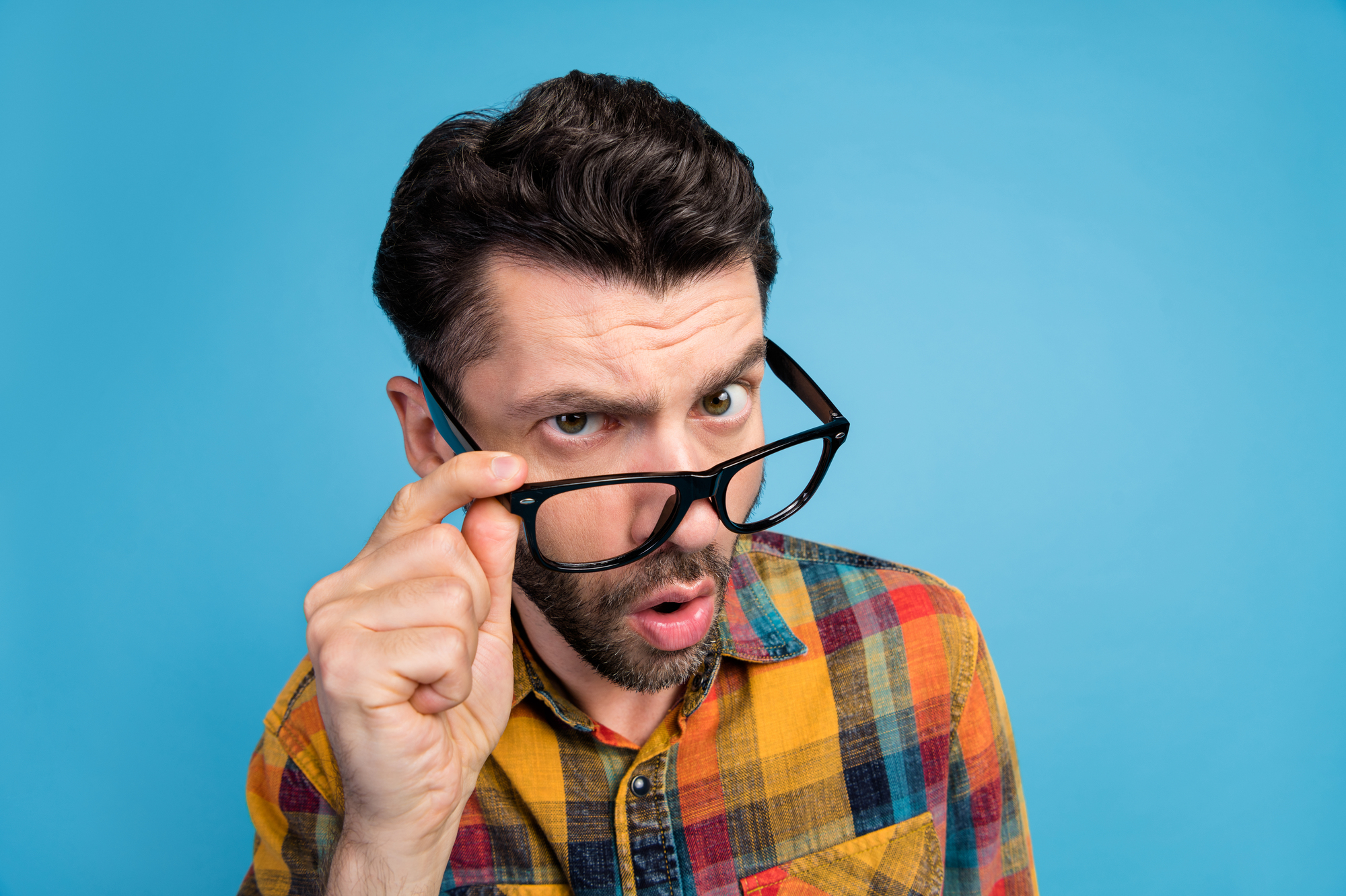 A man with dark hair and a beard is seen against a blue background, wearing a checked shirt with yellow and red colors. He is holding his black-framed glasses down on his nose and looking directly at the camera with a surprised or curious expression.