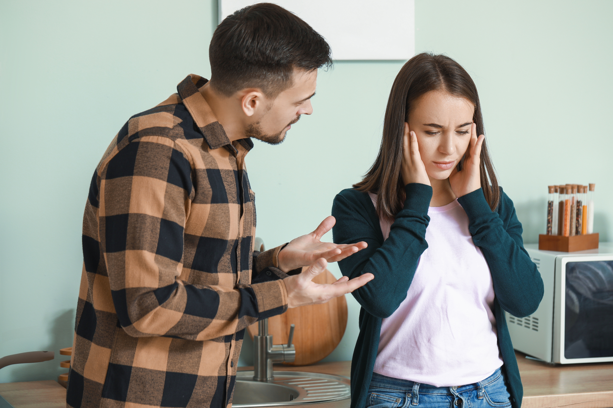 A man with a beard and checkered shirt gestures emphatically while speaking, as a woman with long brown hair and a white shirt covers her ears with a distressed expression. They are in a kitchen with light green walls and appliances in the background.