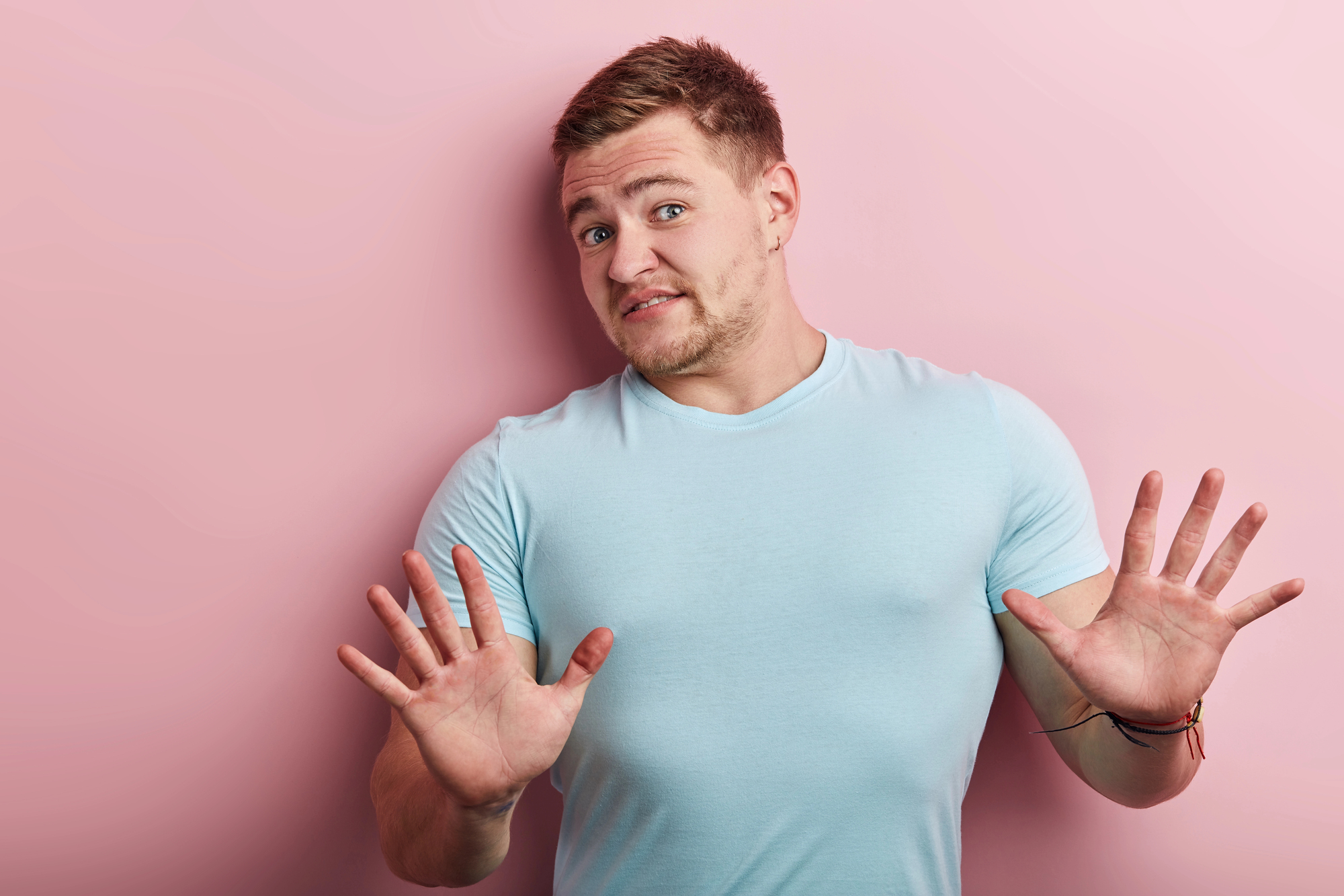 A man wearing a light blue t-shirt stands against a pink background with a hesitant expression on his face. He has his hands raised in front of him, palms facing outward, as if gesturing "stop" or expressing surprise.