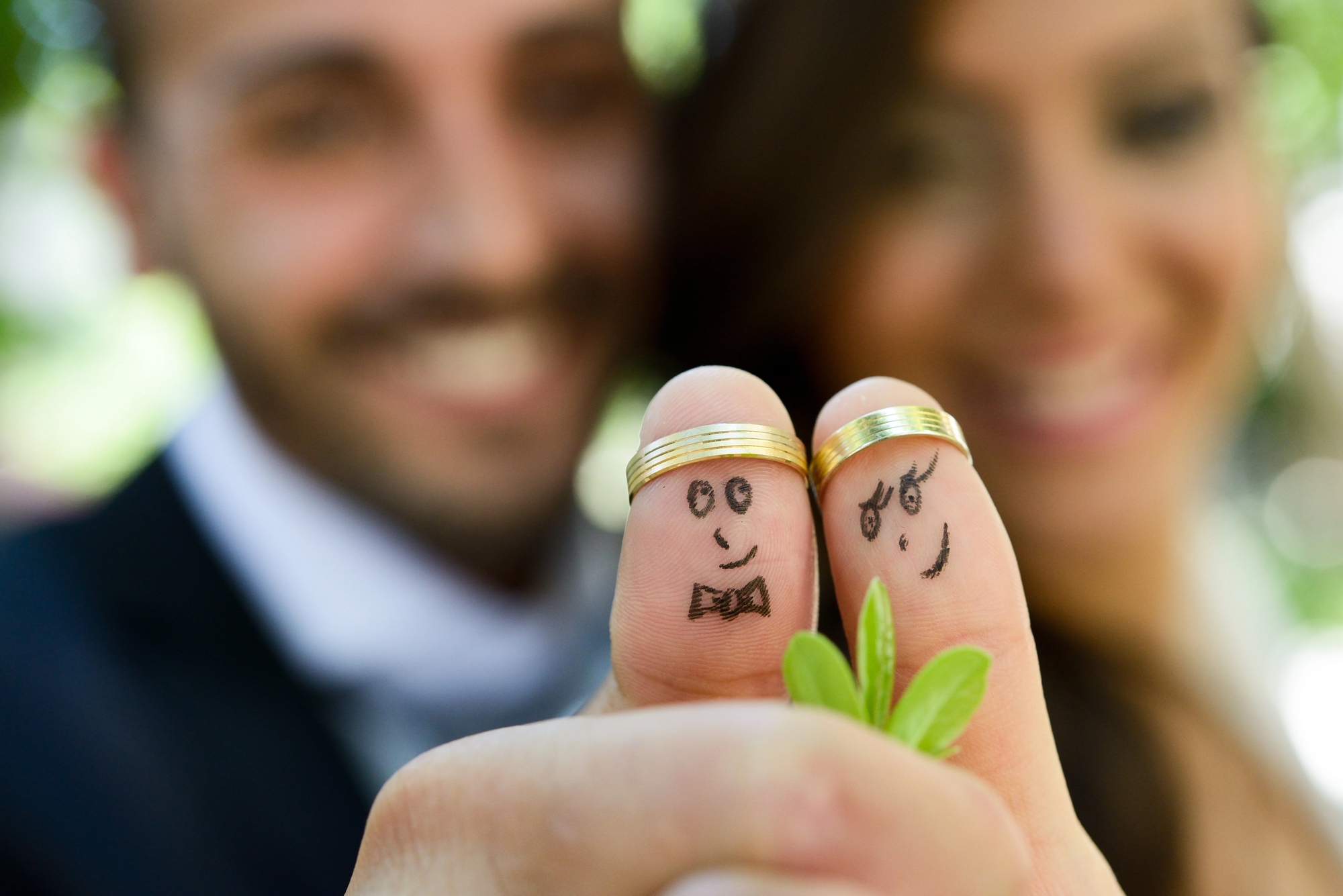 Close-up of two fingers with drawn-on smiling faces, each wearing a gold ring. In the background, a bride and groom slightly out of focus, smiling. The groom's finger has a bow tie drawn on it, while the bride's finger has a simple smile and eyelashes.