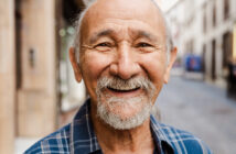 A smiling elderly man with a beard and mustache stands on a city street. He has a bald head and is wearing a blue plaid shirt. The background shows a blurred view of buildings and a pedestrian walkway.