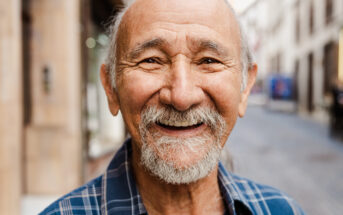 A smiling elderly man with a beard and mustache stands on a city street. He has a bald head and is wearing a blue plaid shirt. The background shows a blurred view of buildings and a pedestrian walkway.