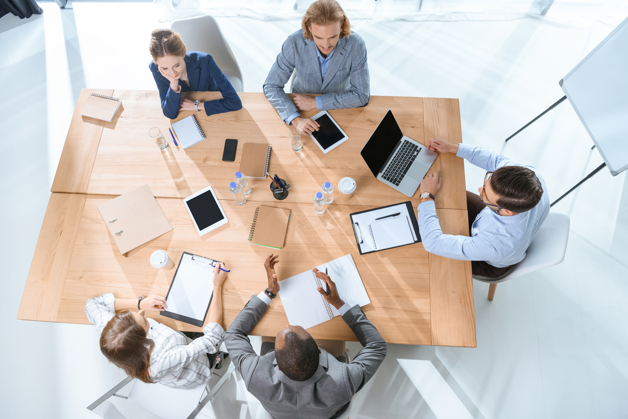 Aerial view of five people sitting around a wooden table engaged in a meeting. They have various devices and documents such as laptops, tablets, notebooks, and sheets of paper. The setting is well-lit, with bright, natural light from large windows.