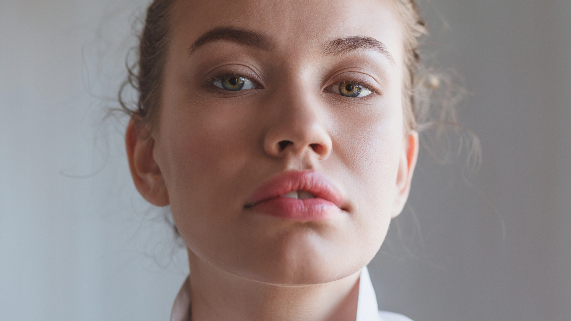 Close-up of a person with light skin, light brown hair, and hazel eyes. They are looking directly at the camera with a neutral expression. The background is blurred, drawing attention to the person's face.