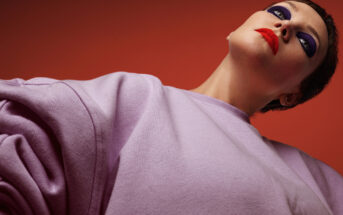 A person with short hair and dramatic makeup, featuring bold red lips and heavy purple eyeshadow, is posed against a red background. They wear a light purple sweater and are viewed from a low angle, looking upwards with a confident, intense expression.