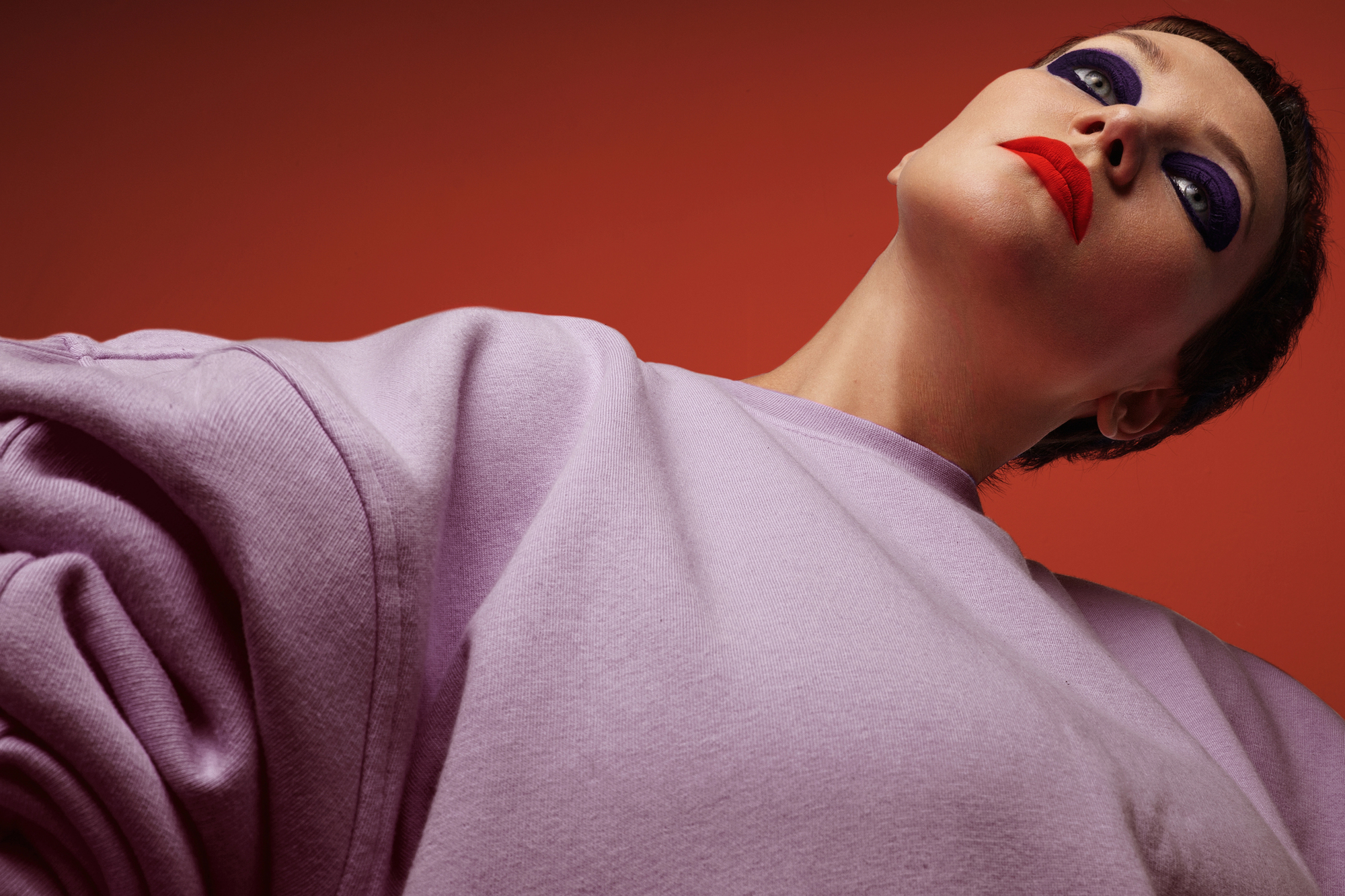 A person with short hair and dramatic makeup, featuring bold red lips and heavy purple eyeshadow, is posed against a red background. They wear a light purple sweater and are viewed from a low angle, looking upwards with a confident, intense expression.