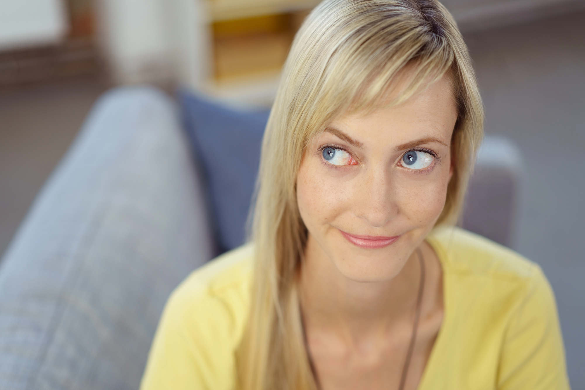 A woman with long blonde hair and blue eyes, wearing a yellow top, is looking slightly to her right with a subtle smile. She is sitting on a light gray couch in a softly lit room. The background is blurred, giving a cozy and relaxed atmosphere.