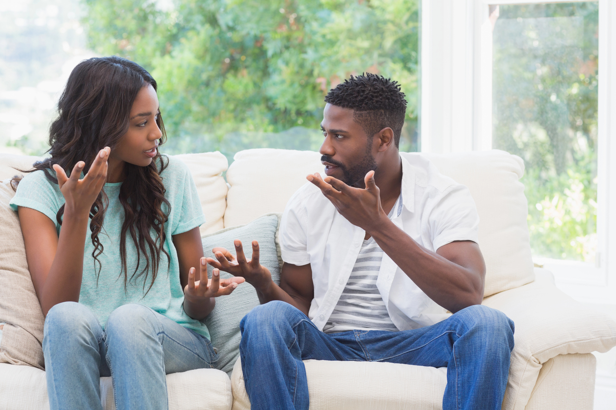 A man and a woman are sitting on a couch engaged in a serious conversation. They are gesturing with their hands, indicating that they are having a disagreement or intense discussion. The background shows a window with a view of green foliage outside.