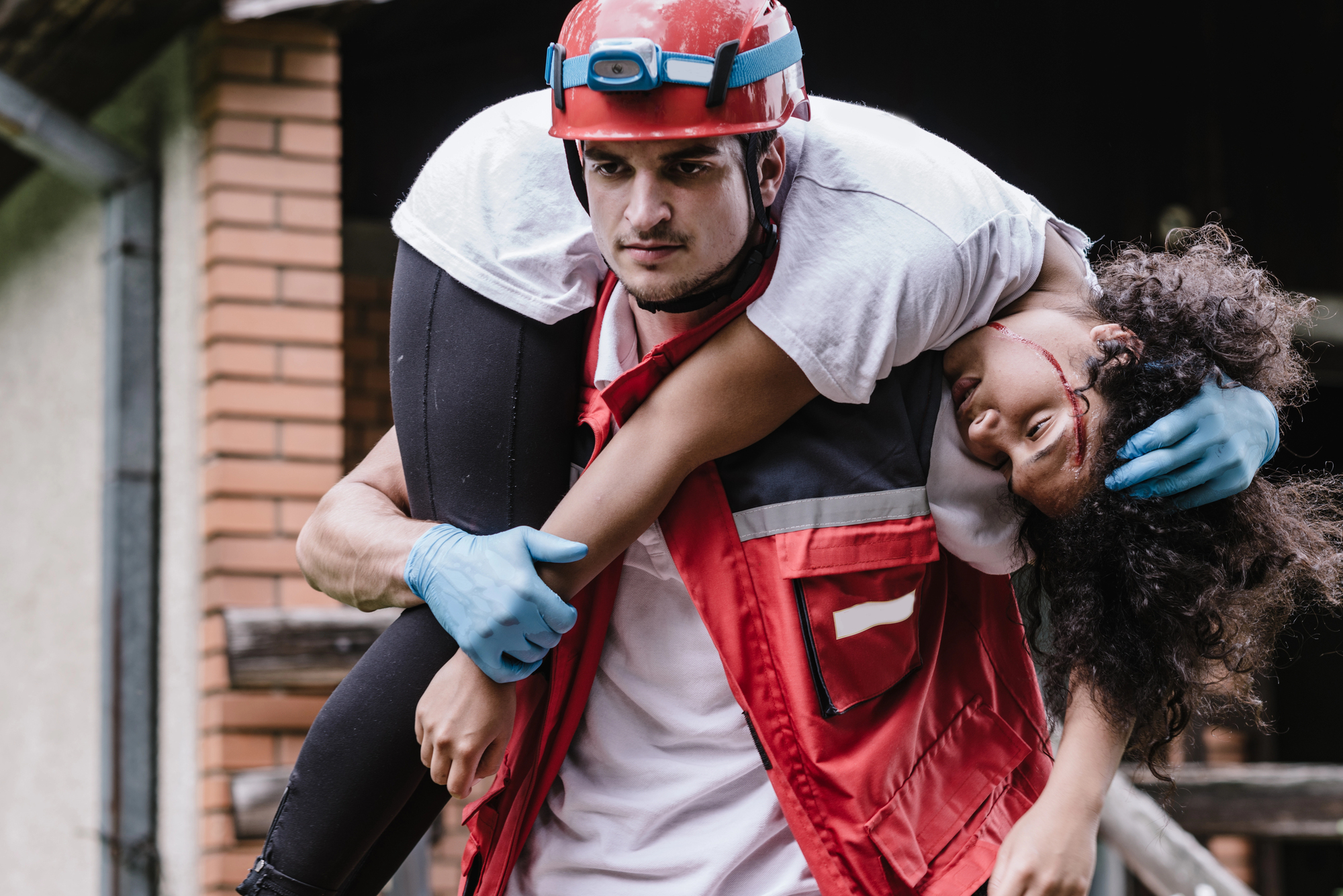 A rescue worker wearing a red helmet and vest carries an injured woman over his shoulder. The woman has a visible cut on her forehead and is unconscious. The background shows a building and wooden structure.