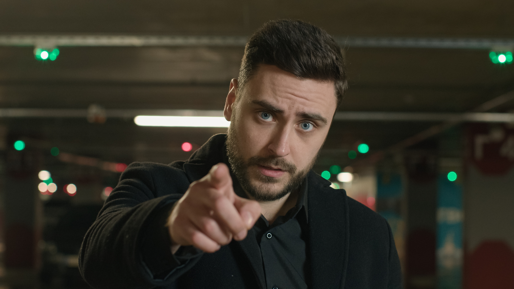 A man with dark hair and beard, wearing a black coat, is standing in a dimly lit indoor area. He is pointing directly at the camera with a serious expression. The background features blurred colored lights and industrial elements.