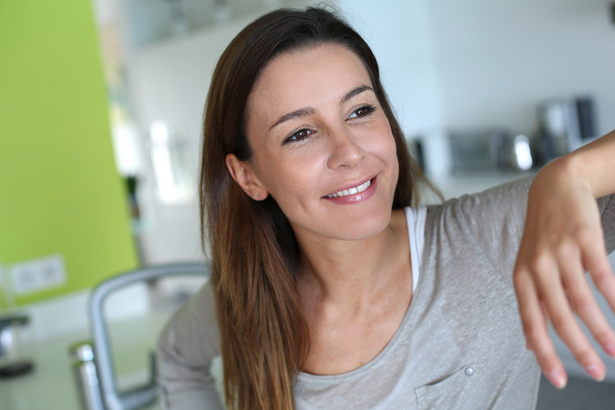 A woman with long, brown hair sits in a bright, modern kitchen. She is wearing a light grey shirt and smiling as she looks off to the side. The background features light green and white walls with kitchen appliances.