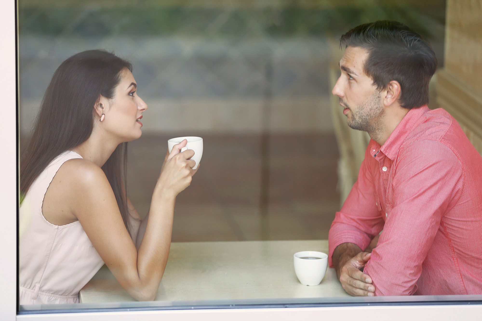 A man and a woman sit at a table in a cafe, holding coffee cups and engaged in conversation. They face each other, with the woman on the left wearing a light-colored top and the man on the right wearing a red shirt. The setting appears cozy and relaxed.