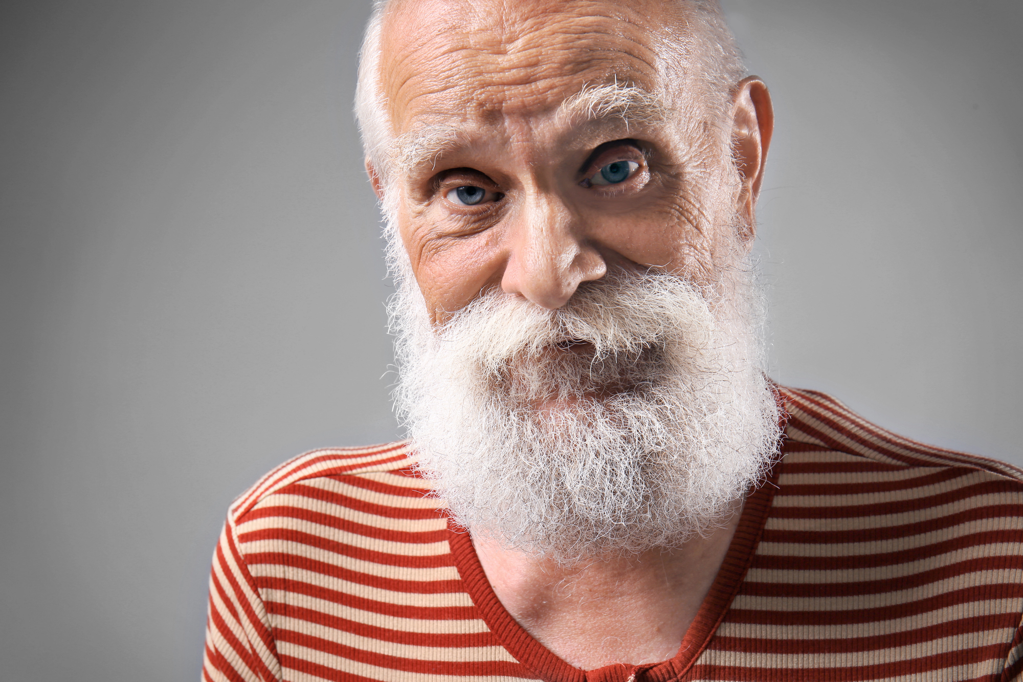 An elderly person with a white beard and mustache smiles warmly at the camera. They are wearing a red and white striped sweater. The background is plain and gray, highlighting the person as the focal point of the image.