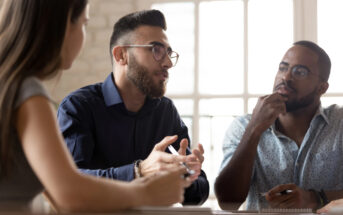 Three people are engaged in a serious discussion around a table. The person in the middle, wearing glasses and a dark shirt, is speaking while gesturing with his hands. The other two individuals, one on each side, are attentively listening. They are in a well-lit room.