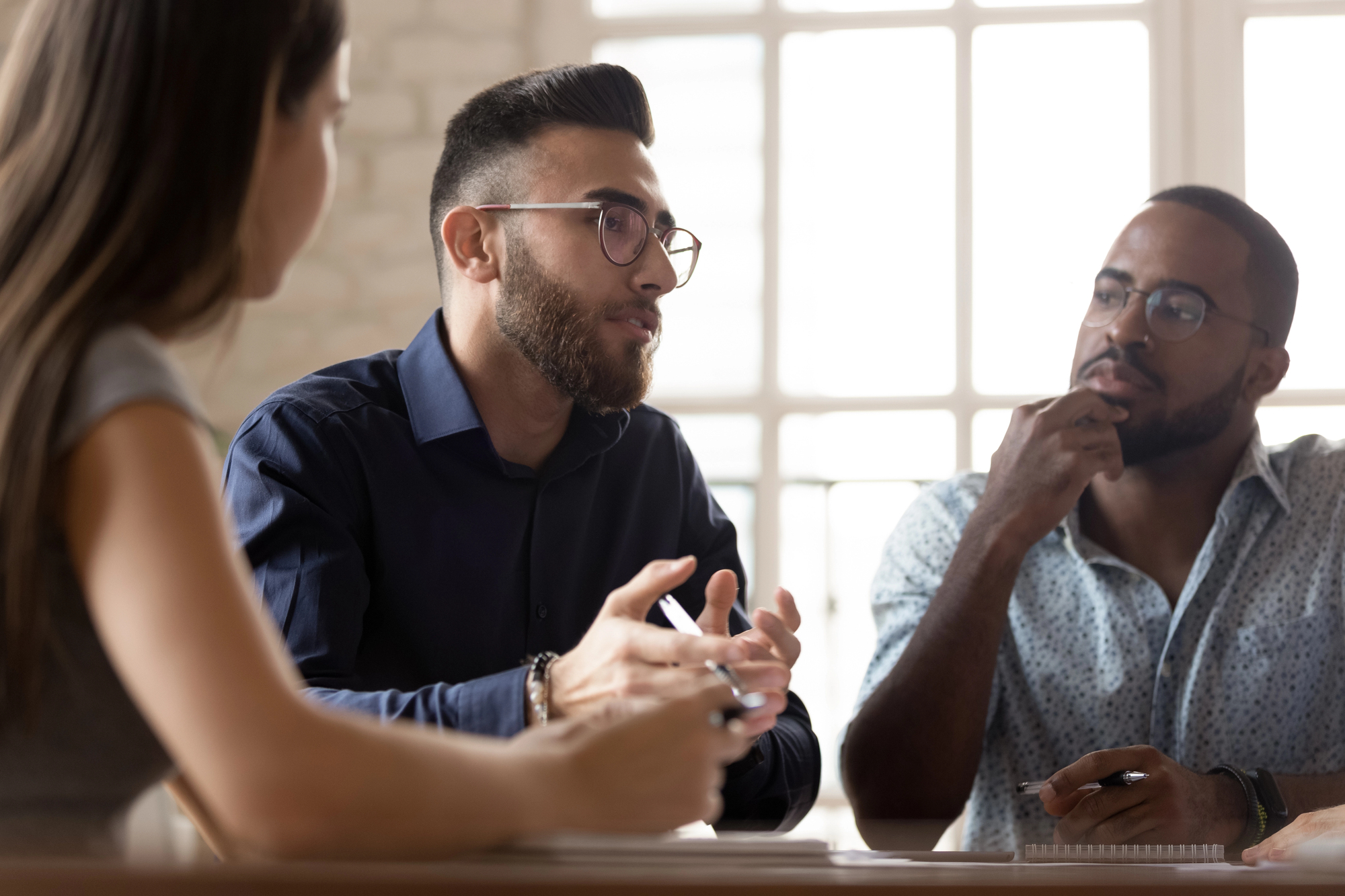 Three people are engaged in a serious discussion around a table. The person in the middle, wearing glasses and a dark shirt, is speaking while gesturing with his hands. The other two individuals, one on each side, are attentively listening. They are in a well-lit room.